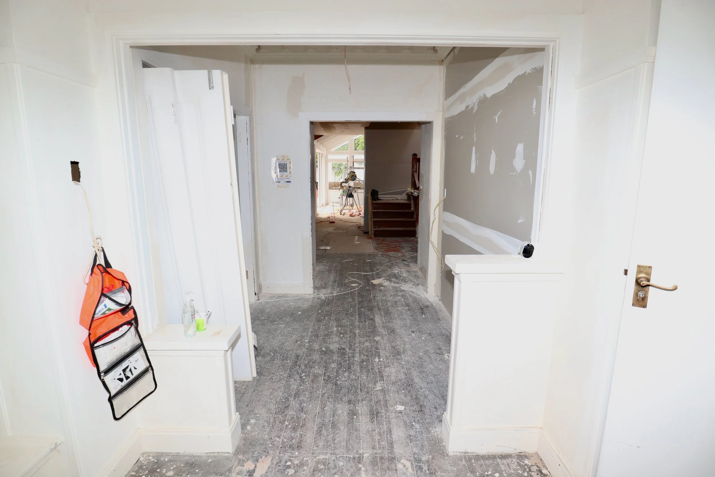 Interior of a house under renovation, with unfinished walls, dusty floor, and construction tools visible.