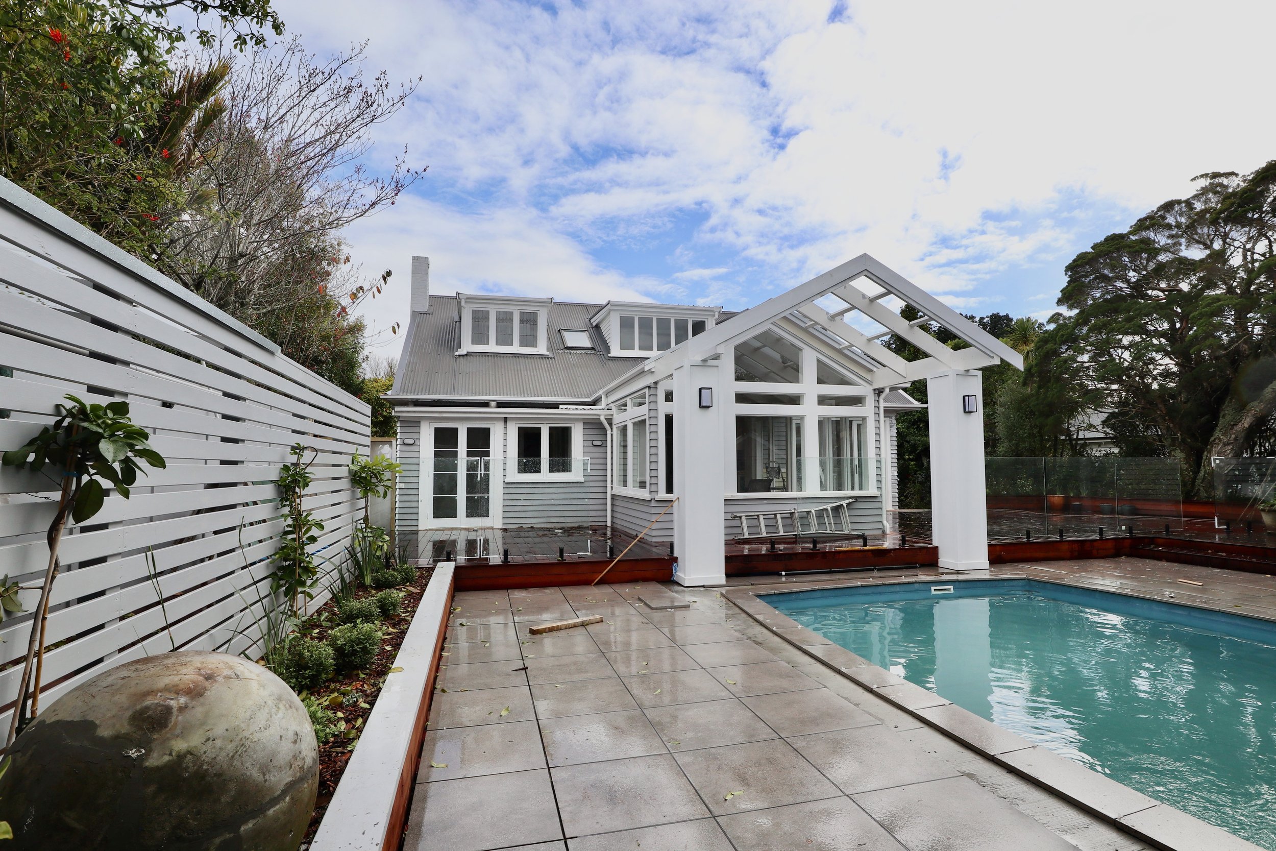 Backyard with a swimming pool, a white house with a glass porch, and a gray roof, surrounded by a white fence and trees, under a partly cloudy sky.