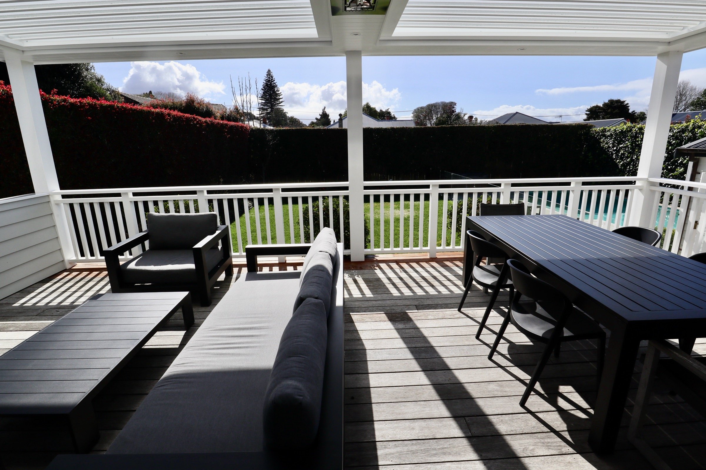 View of a covered outdoor balcony with seating, a wooden table and chairs, a cushioned outdoor sofa, and a view of a green backyard with hedges and houses in the background.