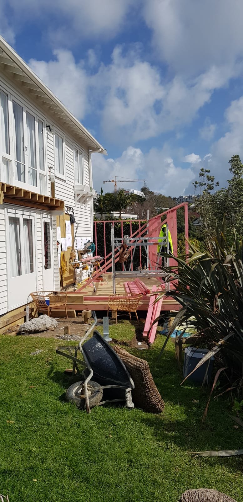 A backyard with a house on the left and a partially constructed deck with pink framing. Construction tools, a wheelbarrow, and outdoor furniture are visible, along with a grassy area and cloudy sky.