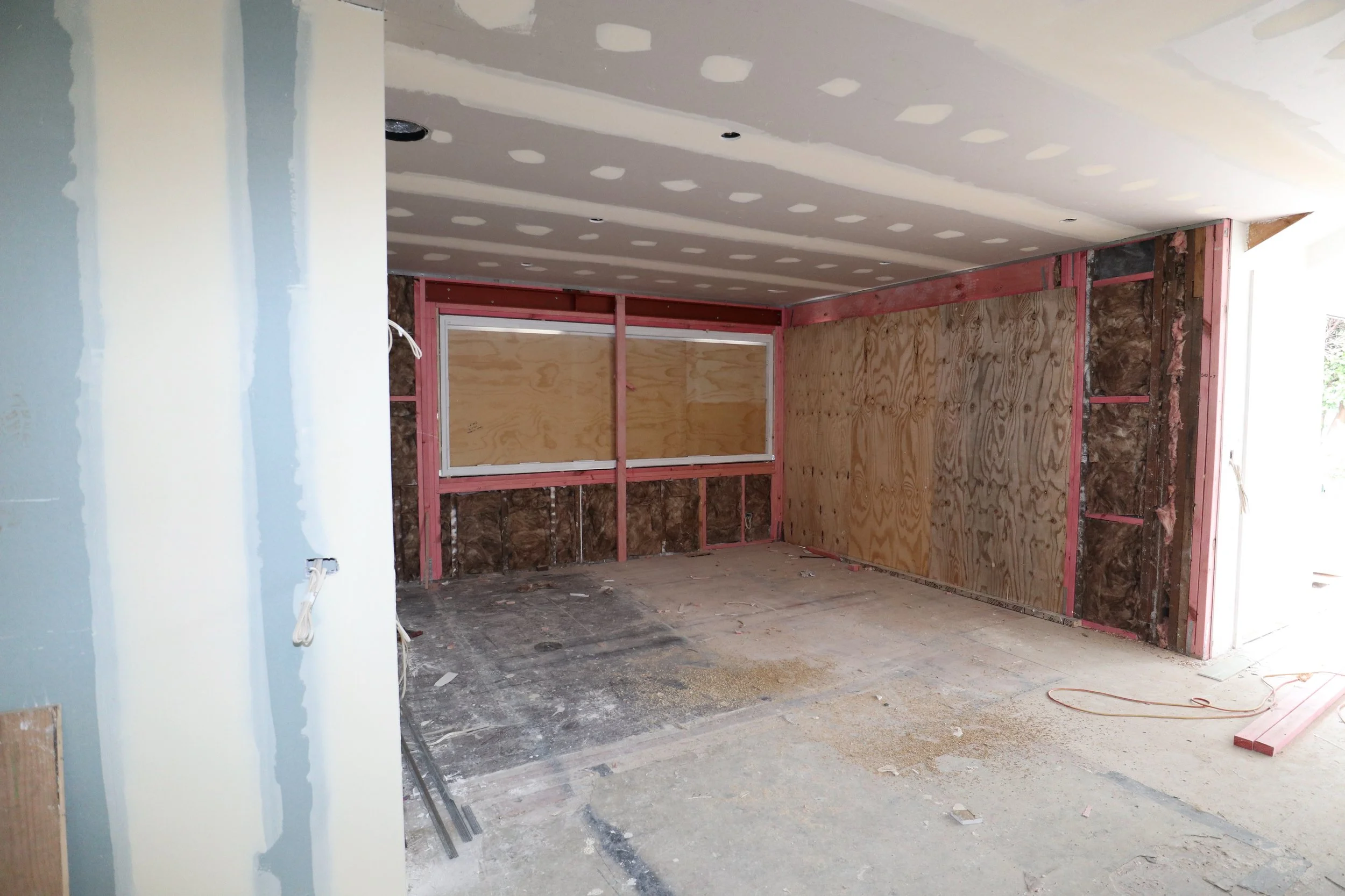Interior view of a room under construction with drywall on the ceiling, partially finished walls with plywood panels, and construction materials on the floor.