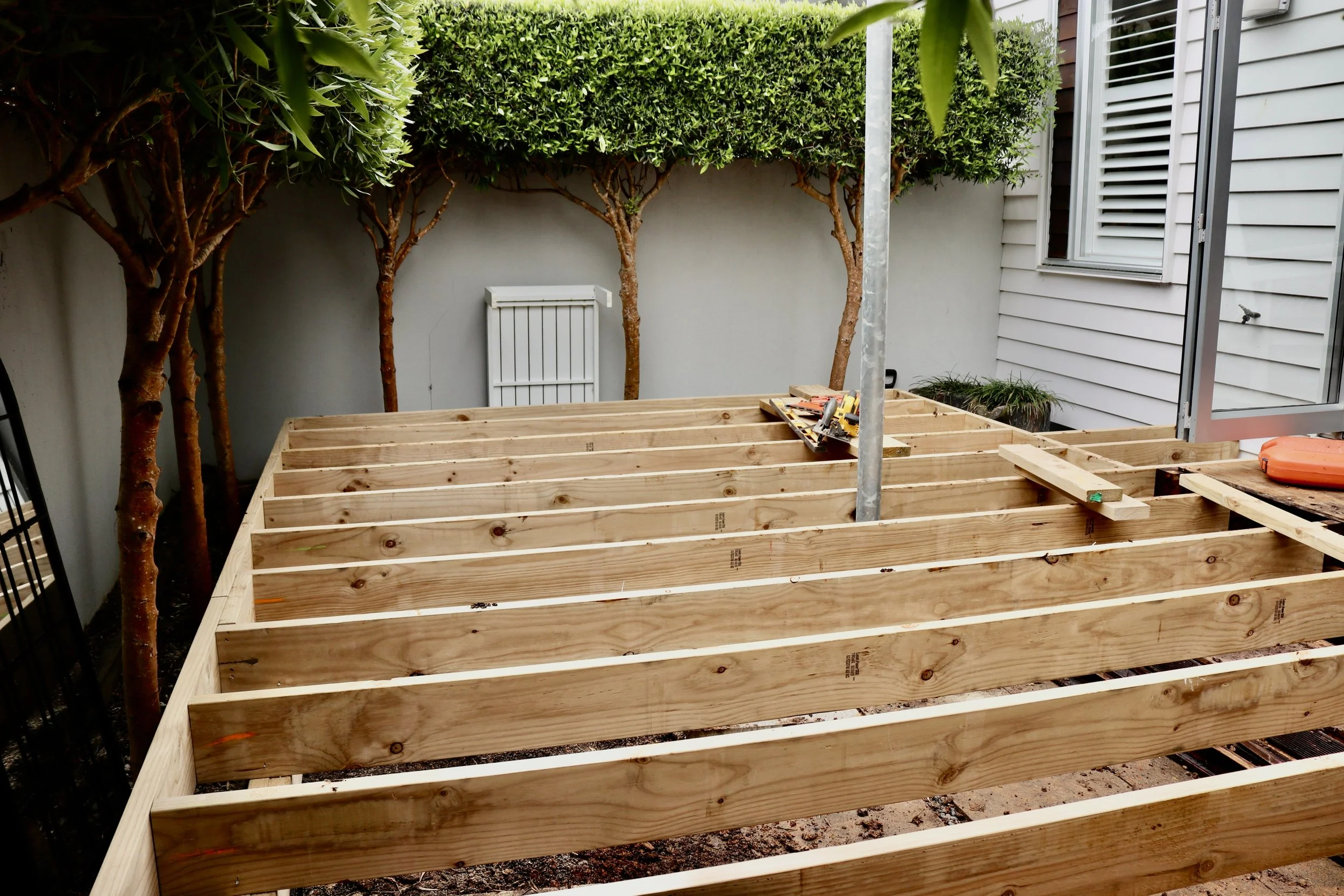 A wooden deck frame under construction in a backyard with trees, a grey wall, and a house with white siding and a sliding glass door.