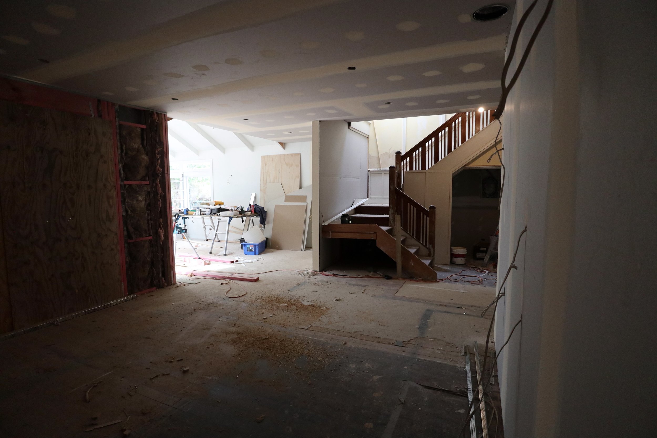 Interior of a house under construction with drywall being installed, construction tools and materials, and a staircase with wooden railings.