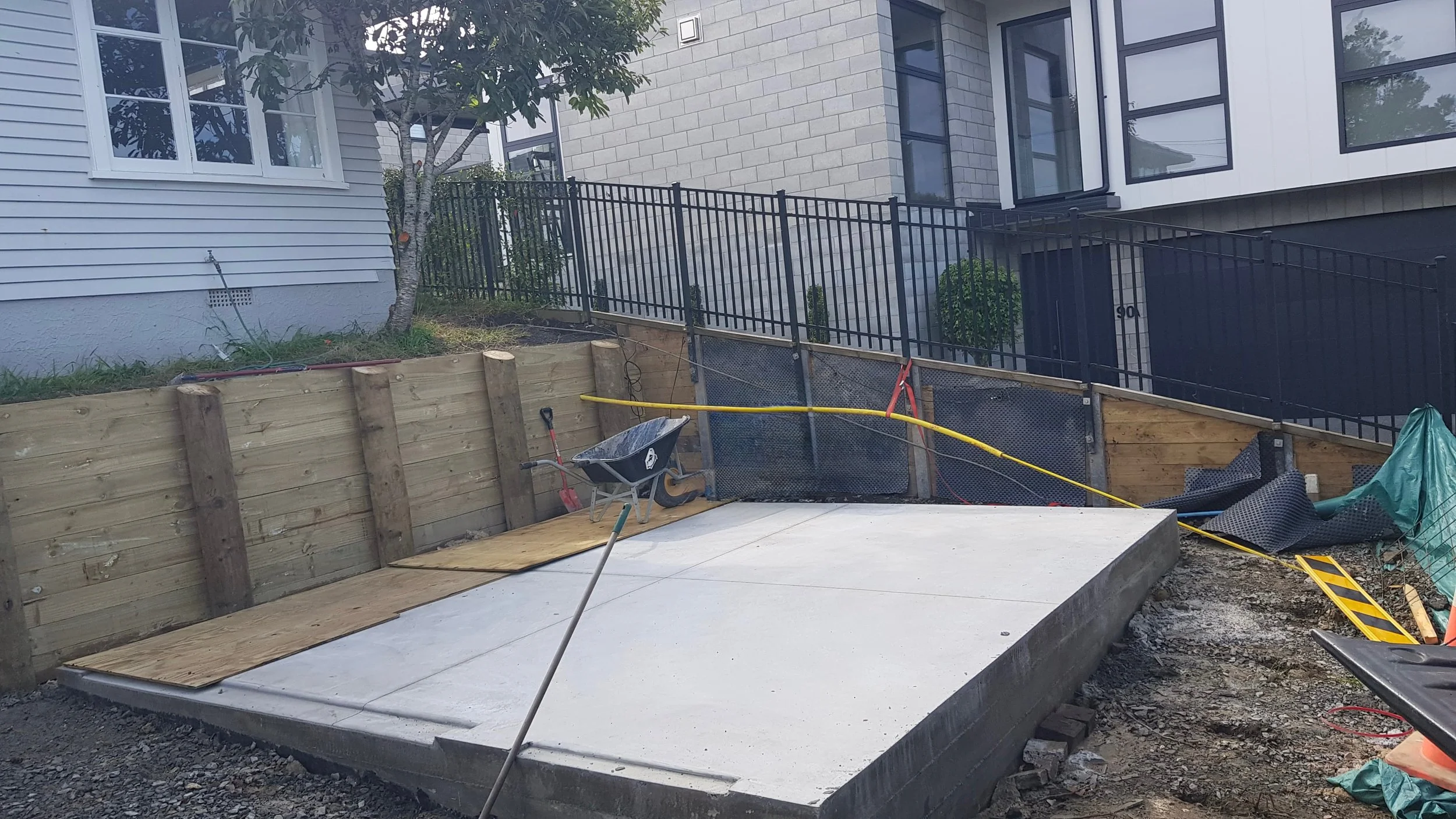 Construction site with freshly poured concrete slab, wooden retaining wall, wheelbarrow, and construction tools, with modern multi-story residential buildings in the background.