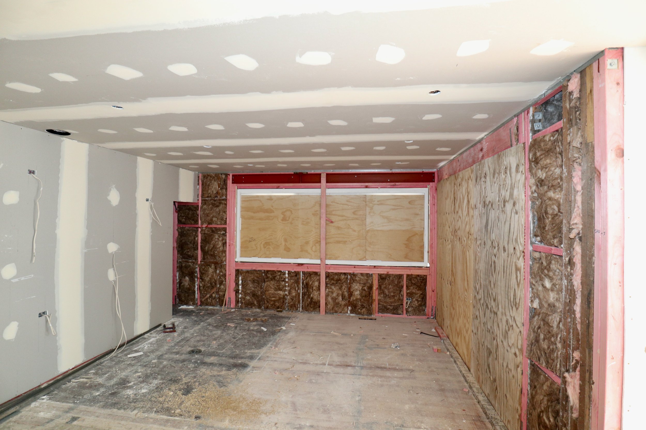 Room under construction with drywall partially installed on ceiling and walls, some areas covered with plywood, and visible insulation, with electrical wires hanging.