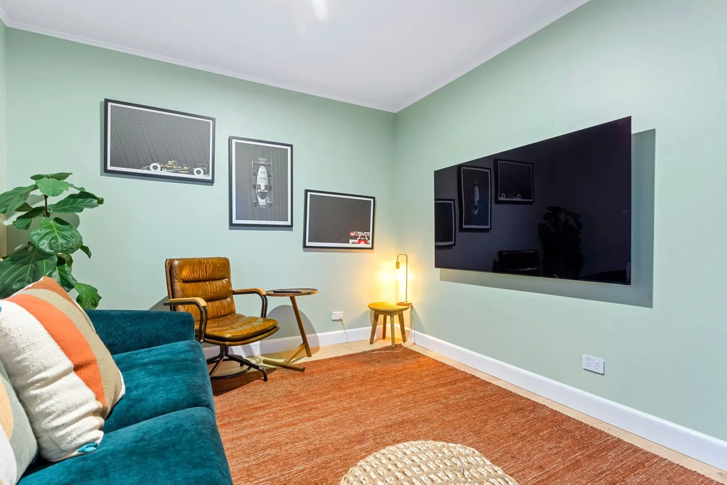 Living room with a teal sofa and a striped cushion, a large potted plant, black-framed artwork on a light green wall, a brown leather armchair, a small wooden side table with a lamp, a large wall-mounted TV, and a rust-colored rug.