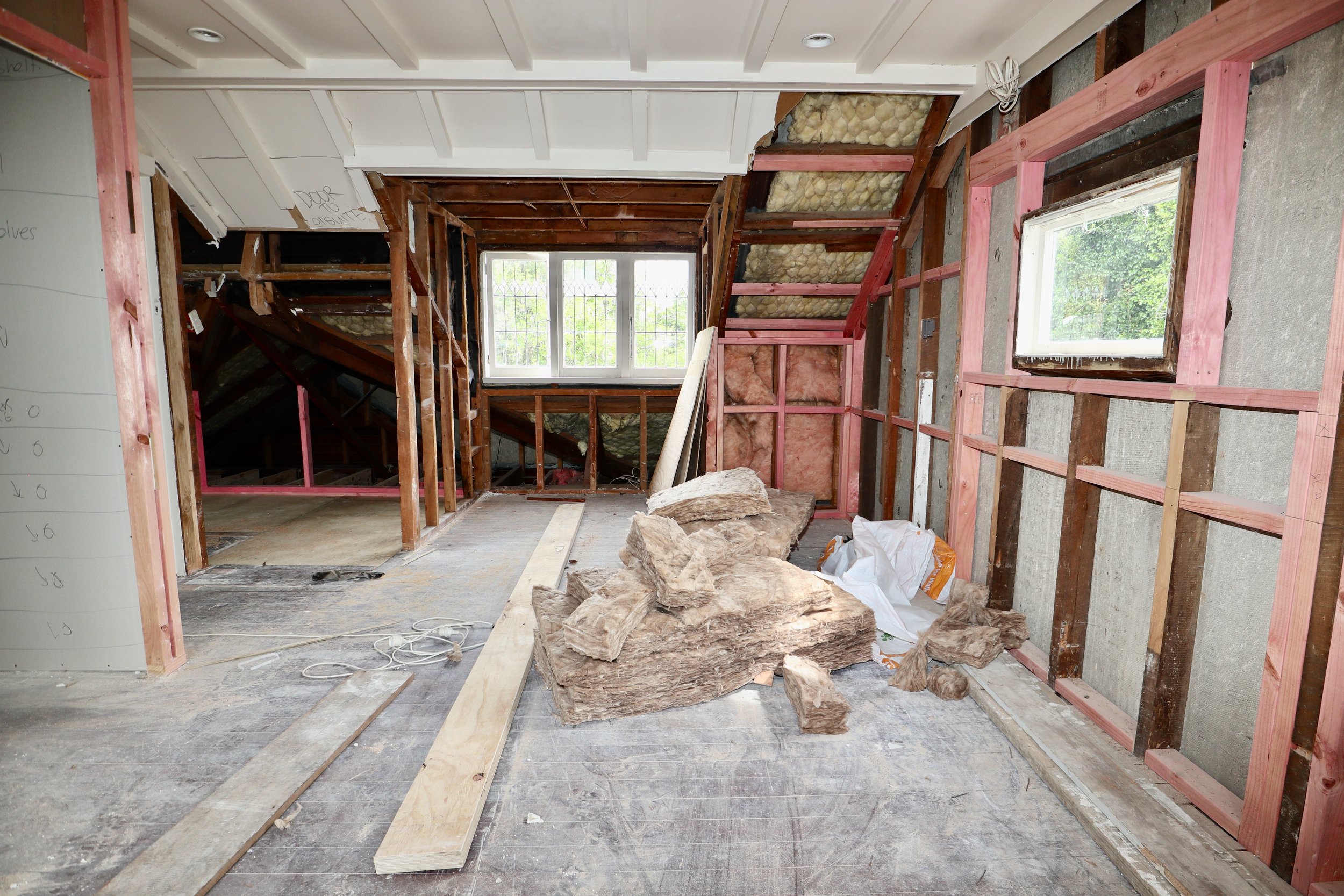 Room under construction with exposed insulation, wooden framing, large rocks on the floor, and a window showing outdoor greenery.