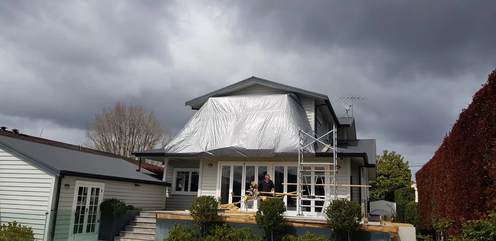 A house under renovation with a construction worker on the deck, a scaffold on the side, and a silver tarp covering part of the roof, with dark storm clouds overhead.