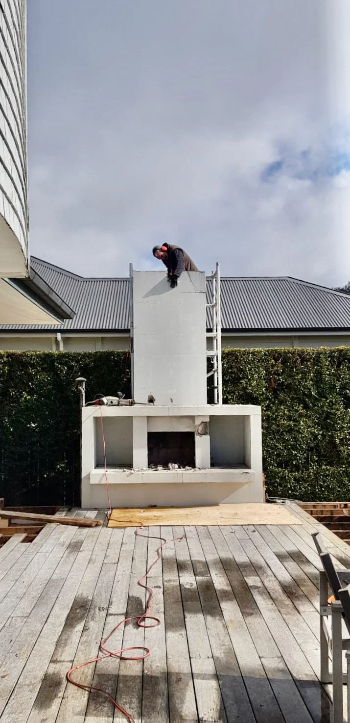 A man on a ladder working on a large outdoor fireplace on a wooden deck, with cloudy sky and house in the background.
