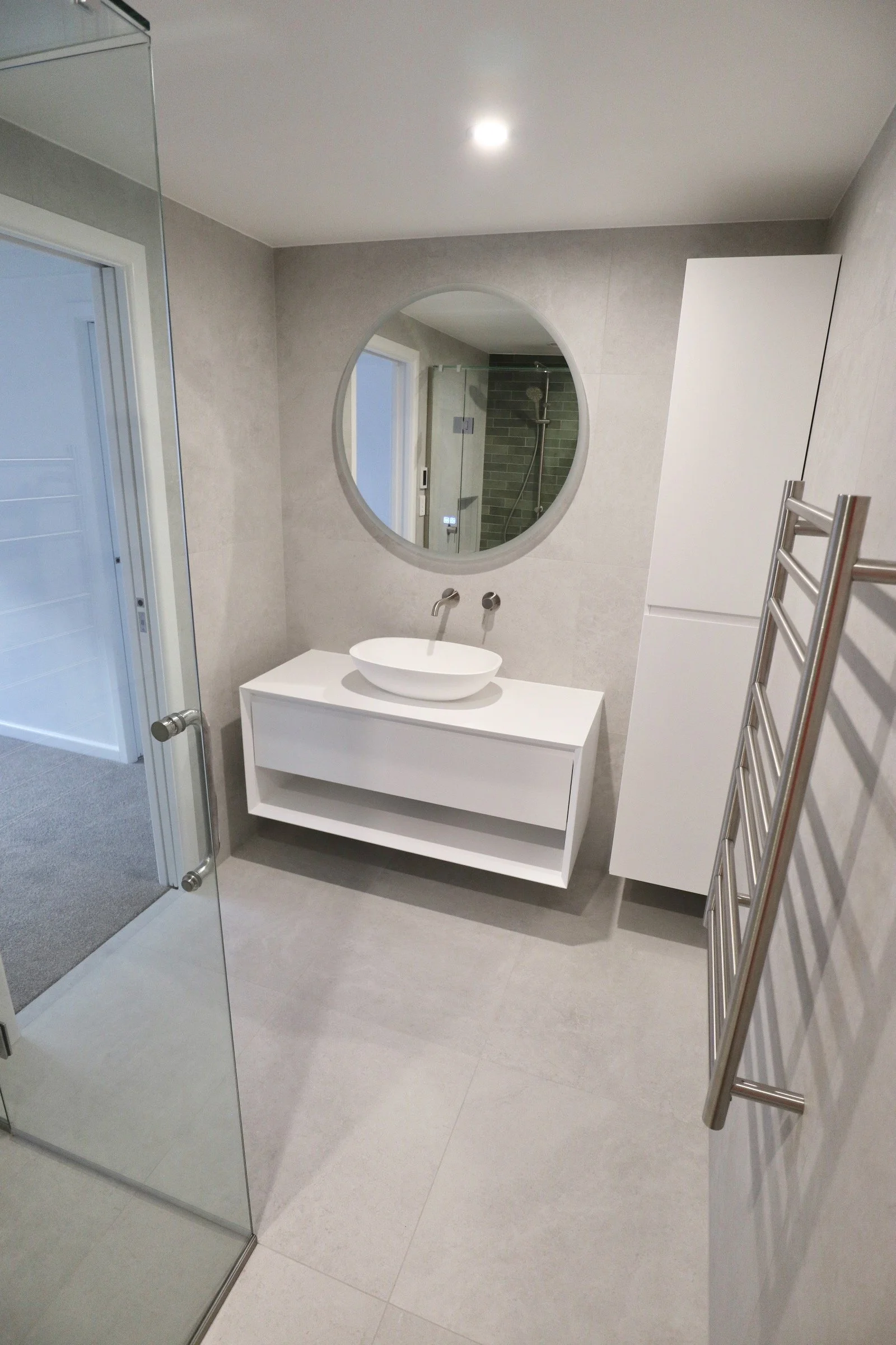Modern bathroom with a floating white vanity, vessel sink, round mirror, towel heater, and walk-in shower with glass door.