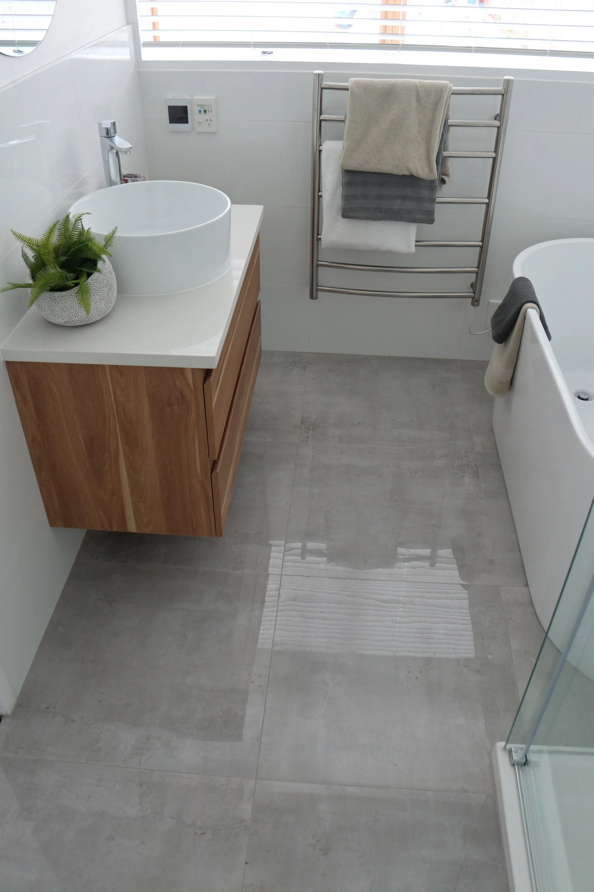 Modern bathroom with gray tiled floor, white walls, a wooden vanity with a round vessel sink, a green potted plant, a heated towel rack with towels, a bathtub with a towel hanging on the side, and a walk-in shower with a glass door.