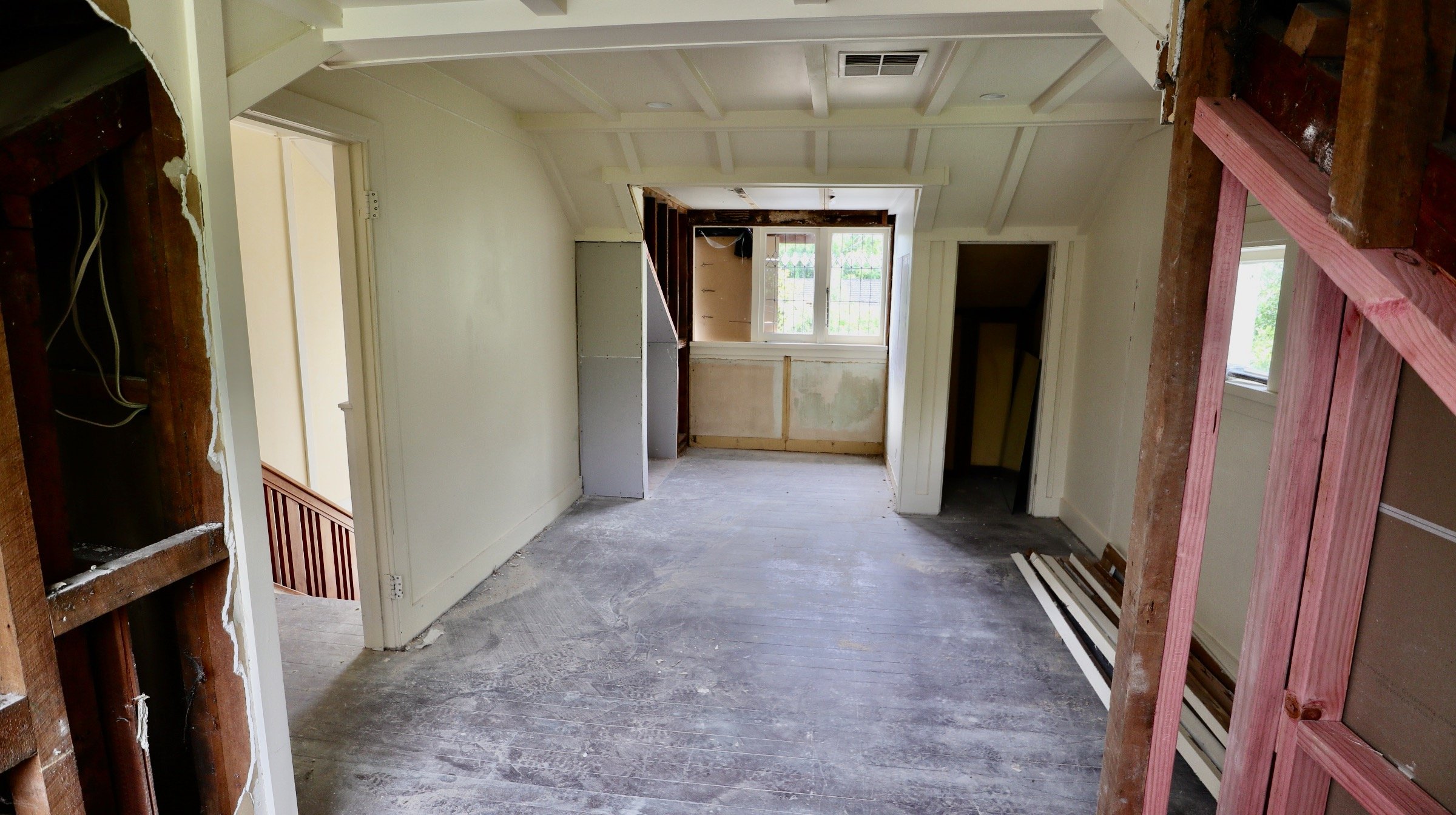 Room under renovation with exposed framing, unfinished flooring, white painted walls, and a window at the far end.