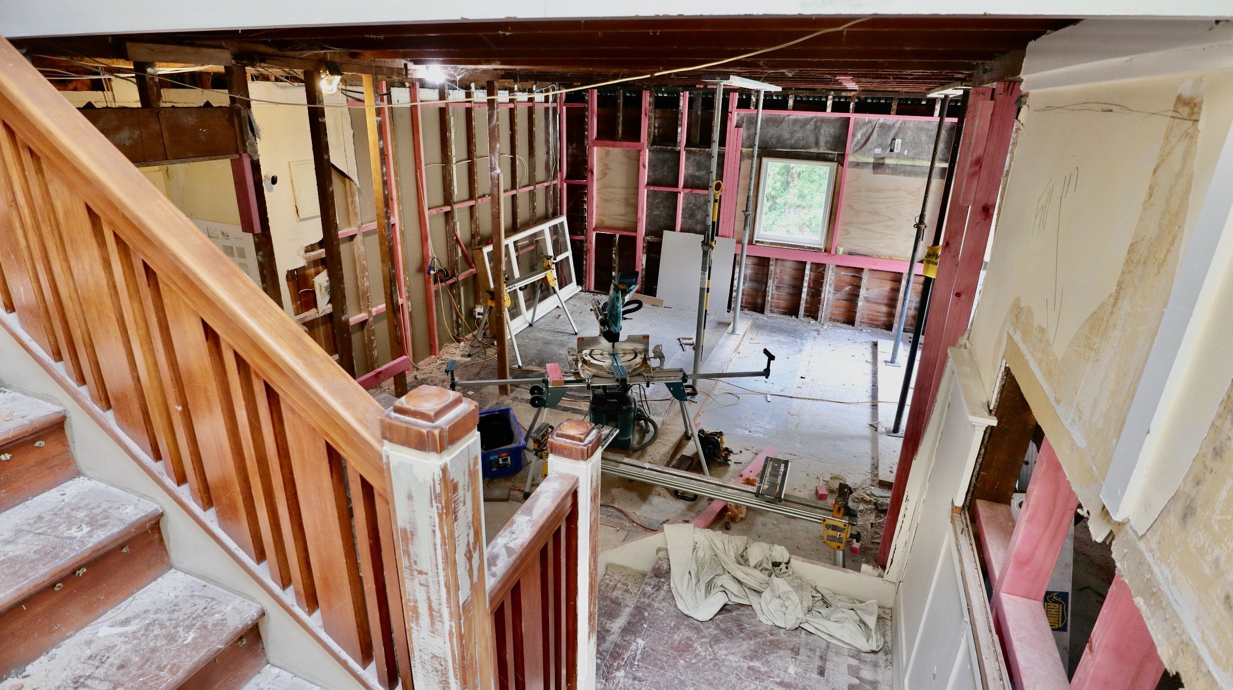Interior of a house under renovation, showing an open space with wooden framing, construction tools, a window, and a staircase with a wooden railing.
