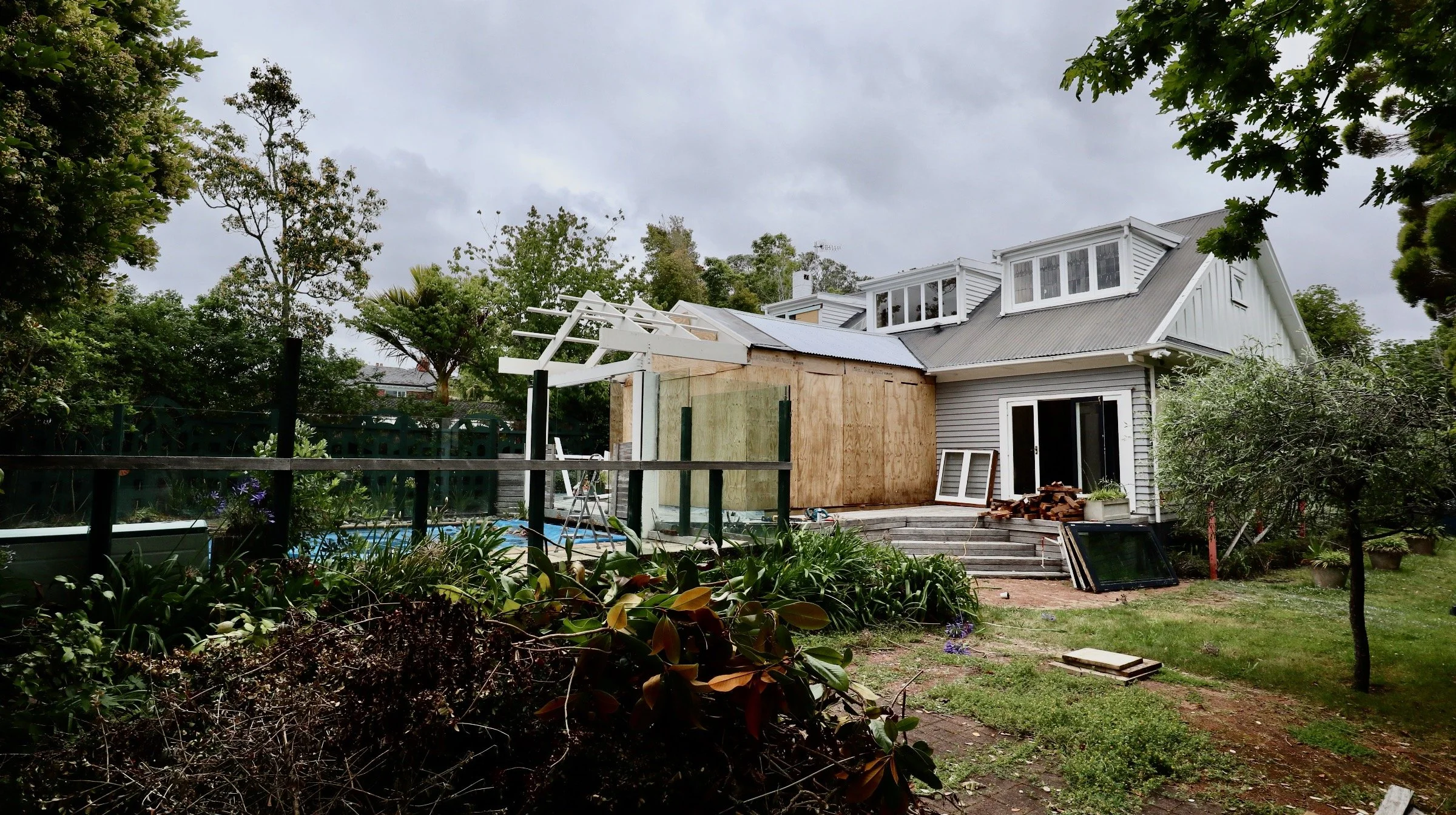 A house under renovation with part of the exterior wall boarded up, construction materials on the steps, and a partially completed pergola in the backyard. Overcast sky with trees surrounding the house.