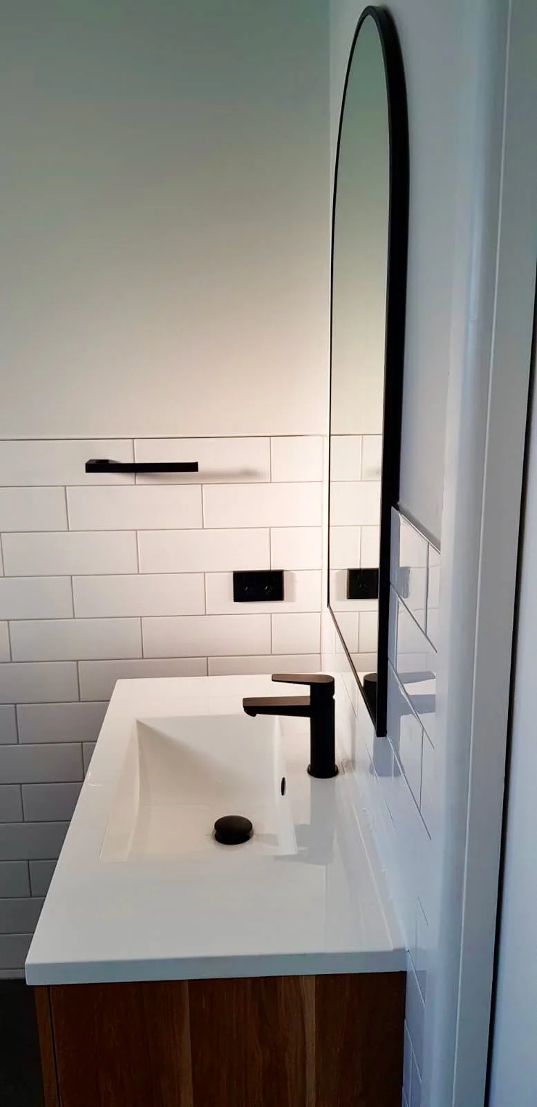A modern bathroom sink with a black faucet, a black soap dish, white tiled wall, and a black oval mirror.