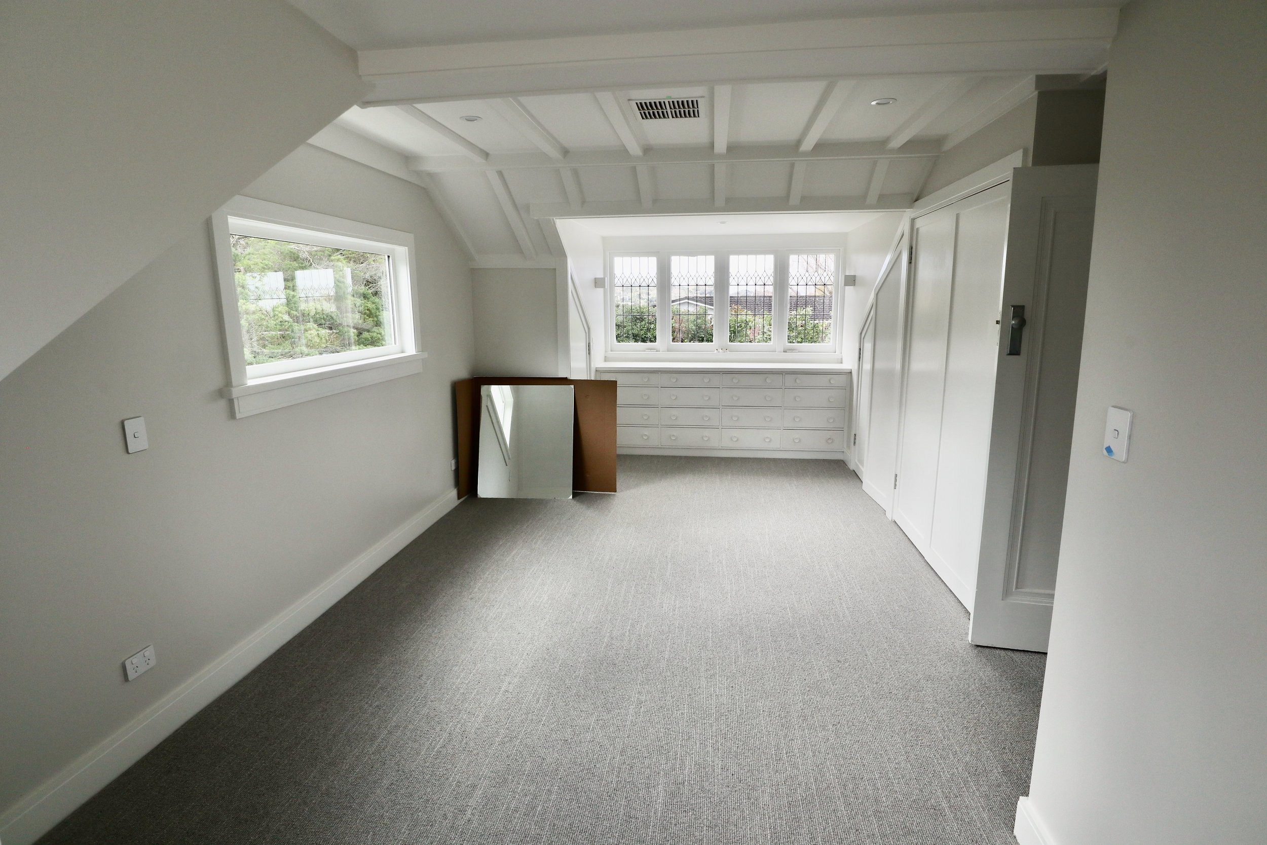 Empty room with white walls, gray carpet, multiple windows, built-in white drawers, and a standing mirror leaning against the wall.