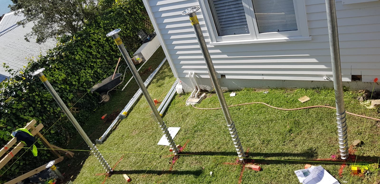 Construction site with four metal poles and yellow string marking layout, house with white siding and window in background, tools and materials scattered on grass