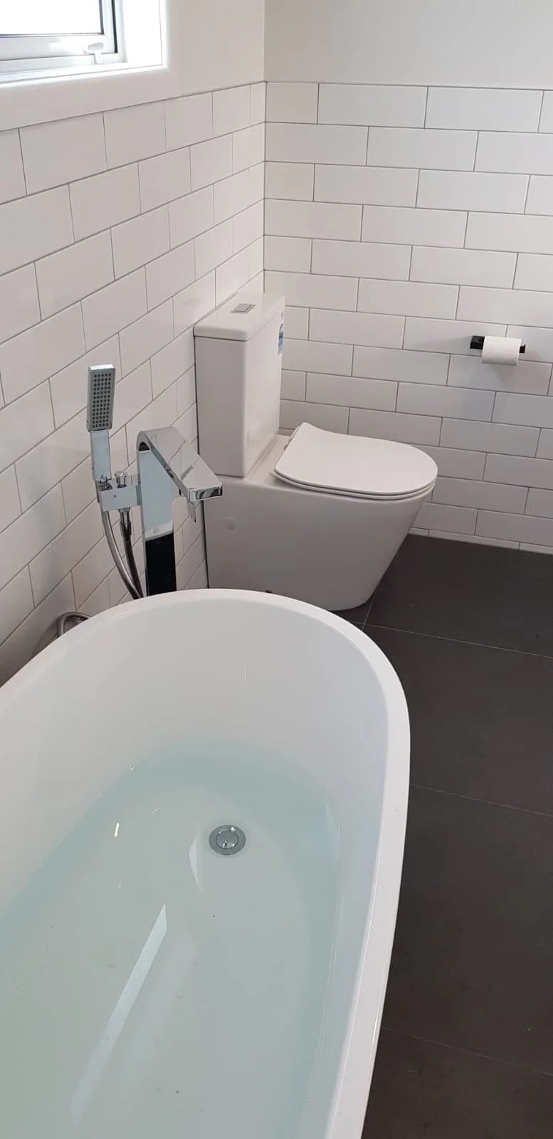 A modern bathroom with a freestanding bathtub filled with water, a wall-mounted faucet, a toilet with a rectangular tank, a wall-mounted toilet paper holder with a roll, white tiled walls, and a small window.