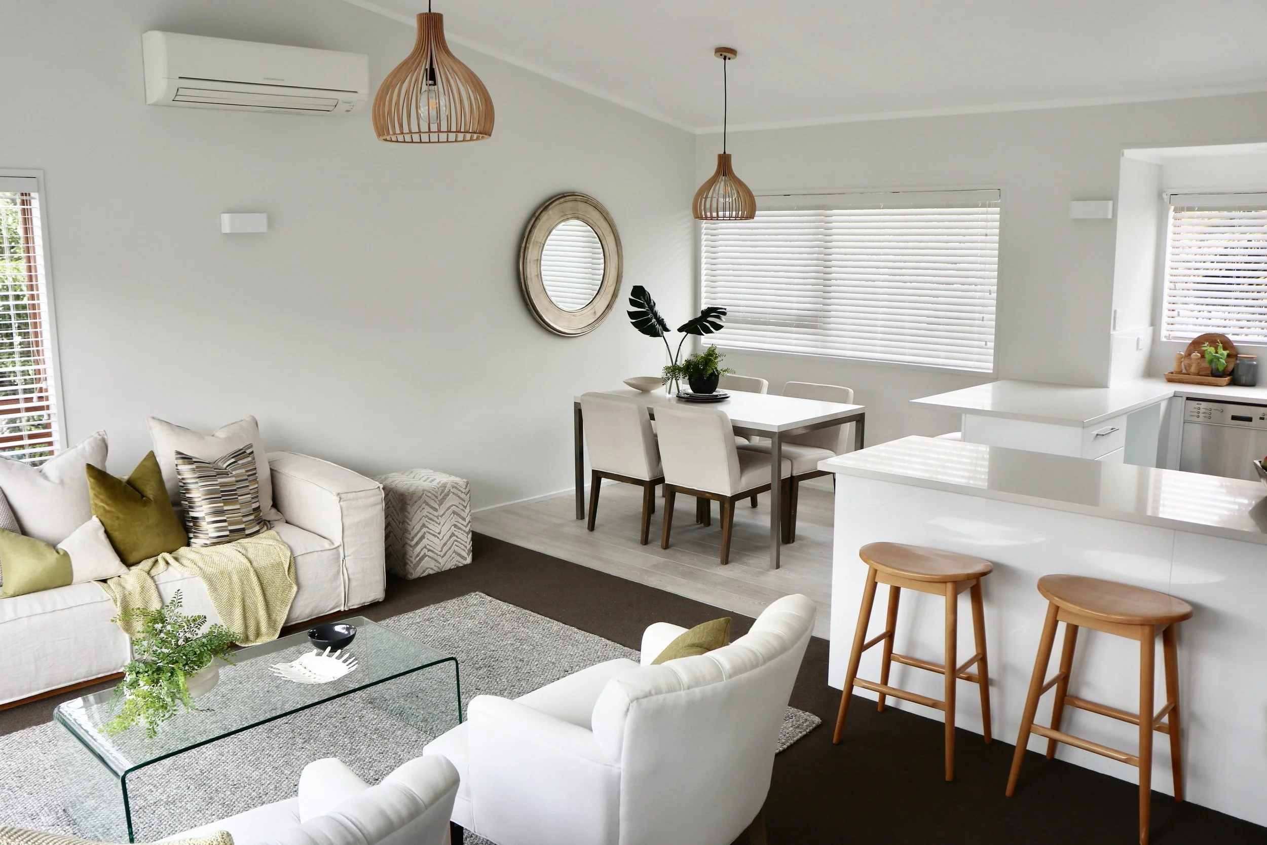 Bright living room with white furniture, a glass coffee table, and a dining area with a white table and chairs, near a white kitchen with wooden bar stools and large windows with blinds.
