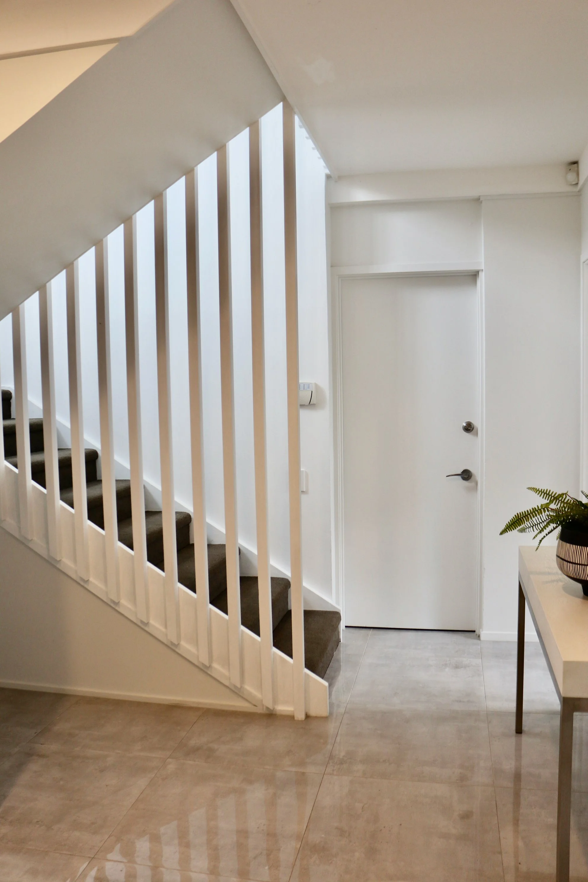 Interior view of a modern home with a staircase, white walls, a white door, beige tiled floor, and a small table with potted plant.