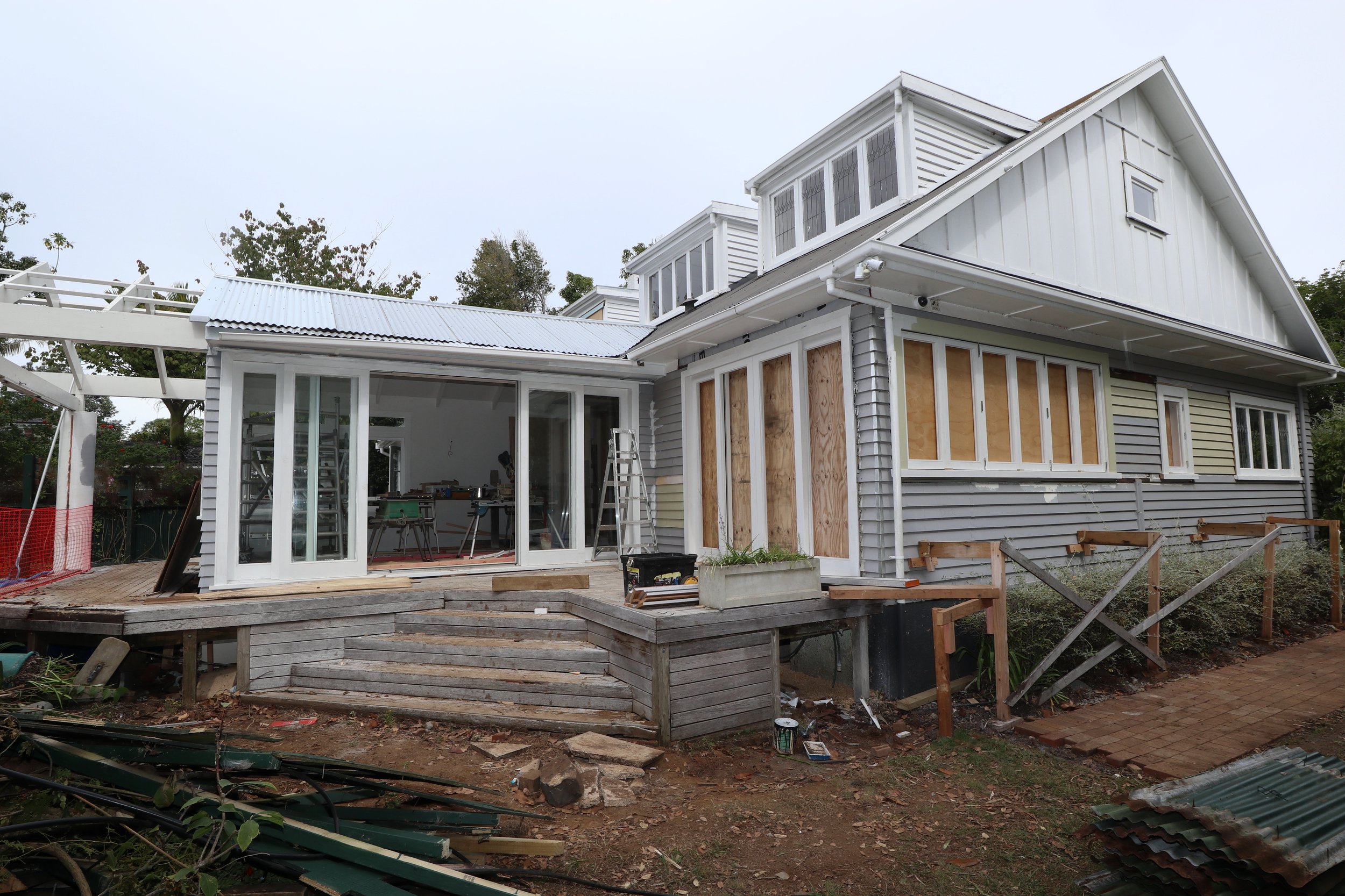 A house under construction with construction materials, tools, and temporary coverings in a backyard, showing partially finished walls and windows.