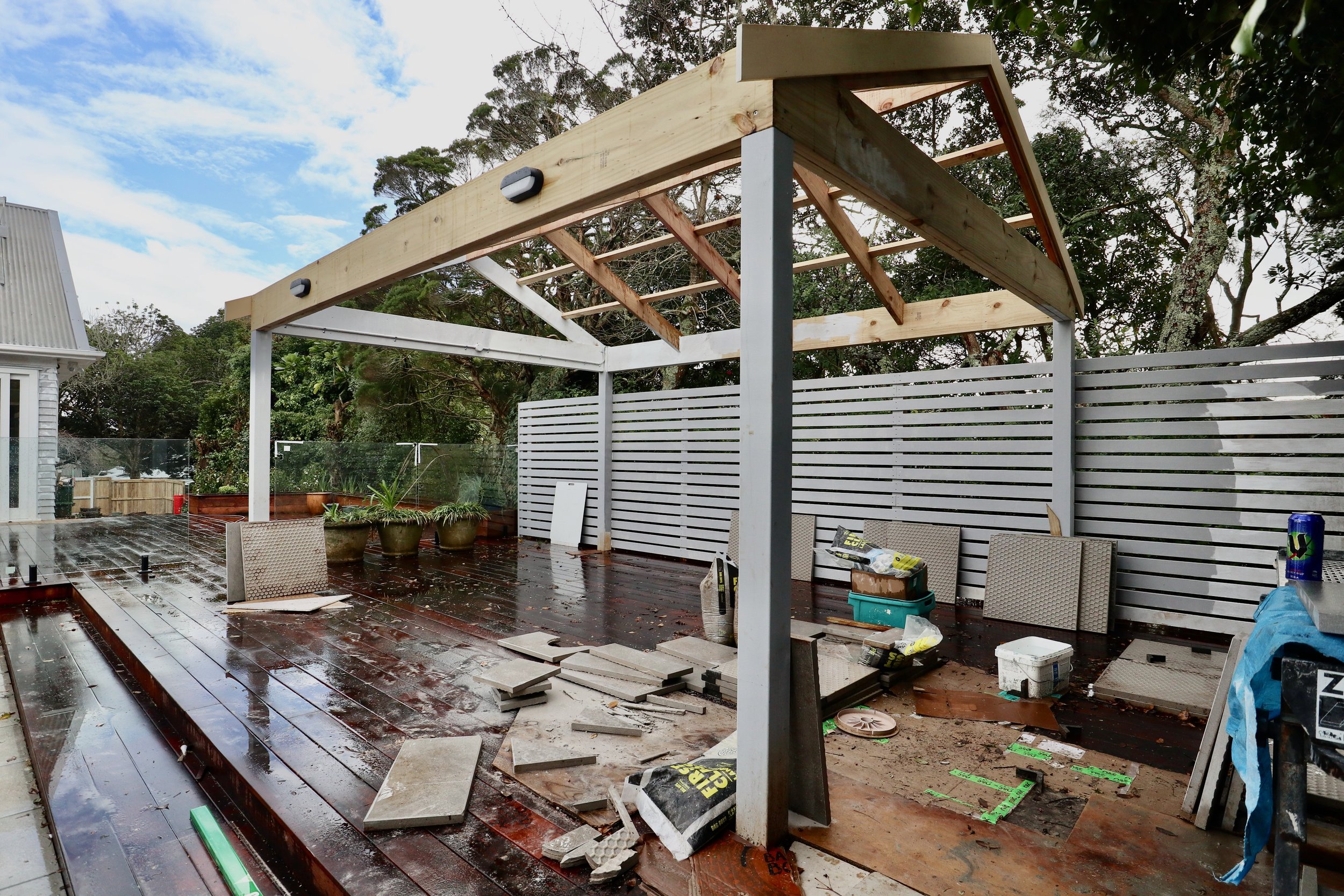 Under construction outdoor patio with a partially built wooden roof structure, potted plants, tools, and construction materials on a wet wooden deck surrounded by a white fence and trees.