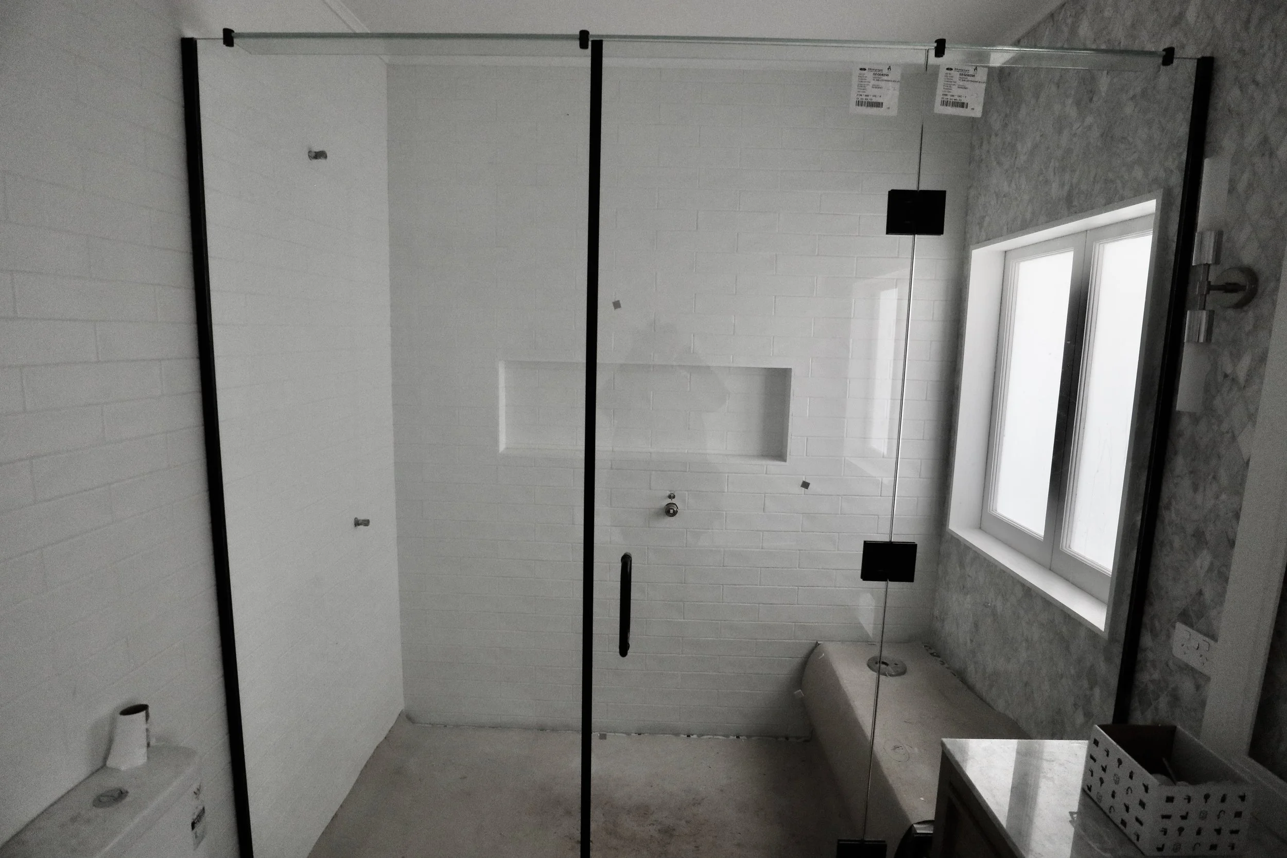An unfinished bathroom with a glass shower door, a white brick wall, and a window on the right side. There are construction materials and tools present.