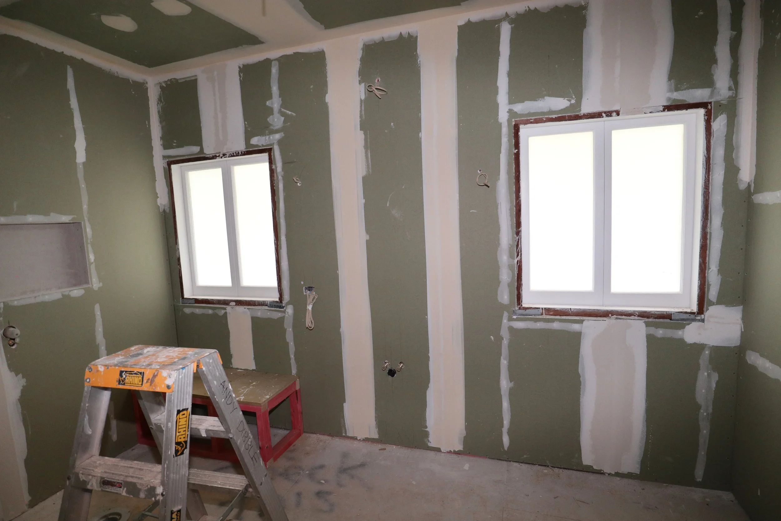 A room under construction with bare drywall and exposed seams, two white-framed windows, and a step ladder in the foreground.
