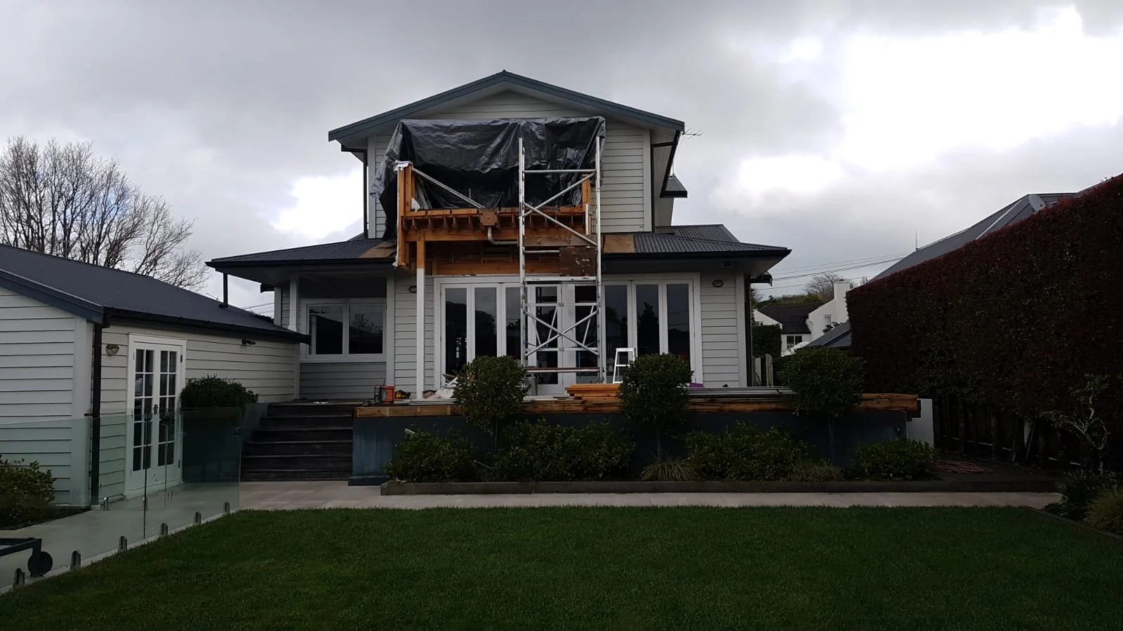 House under construction with scaffolding, partially built deck, and a tarp covering the upper balcony, surrounded by a well-maintained lawn and hedge, with cloudy sky in the background.