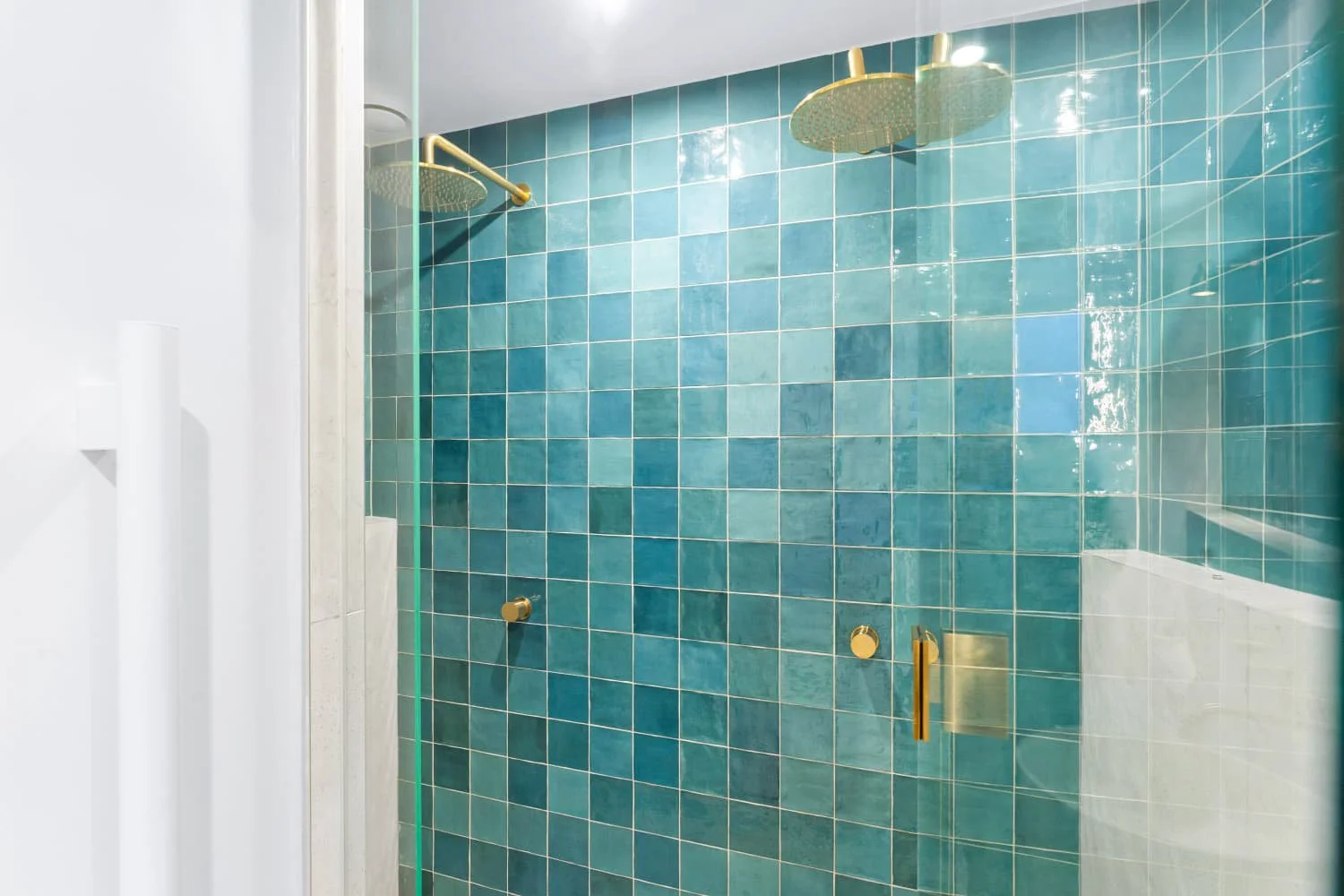 Glass shower enclosure with brass fixtures, featuring two ceiling-mounted rain shower heads and a glass door with brass handle, set against teal tiled wall.
