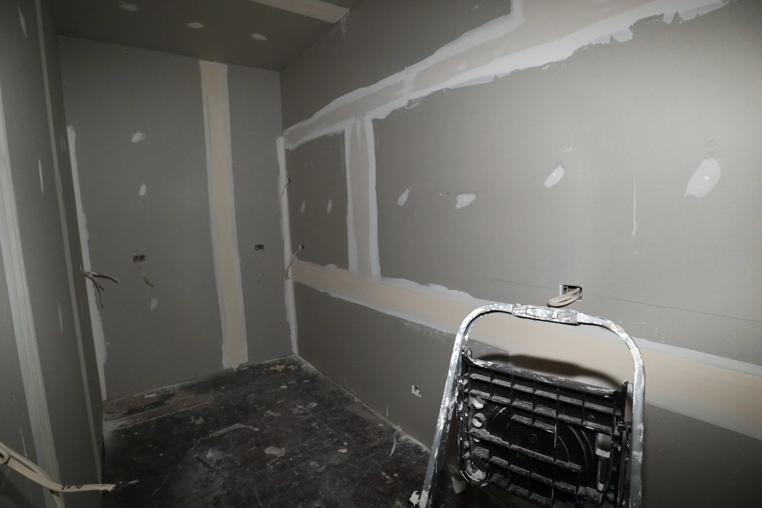 A room under construction with drywall installed, patching and joint compound applied, and electrical outlets with wires hanging. There is a step ladder in the foreground and a black floor with debris.
