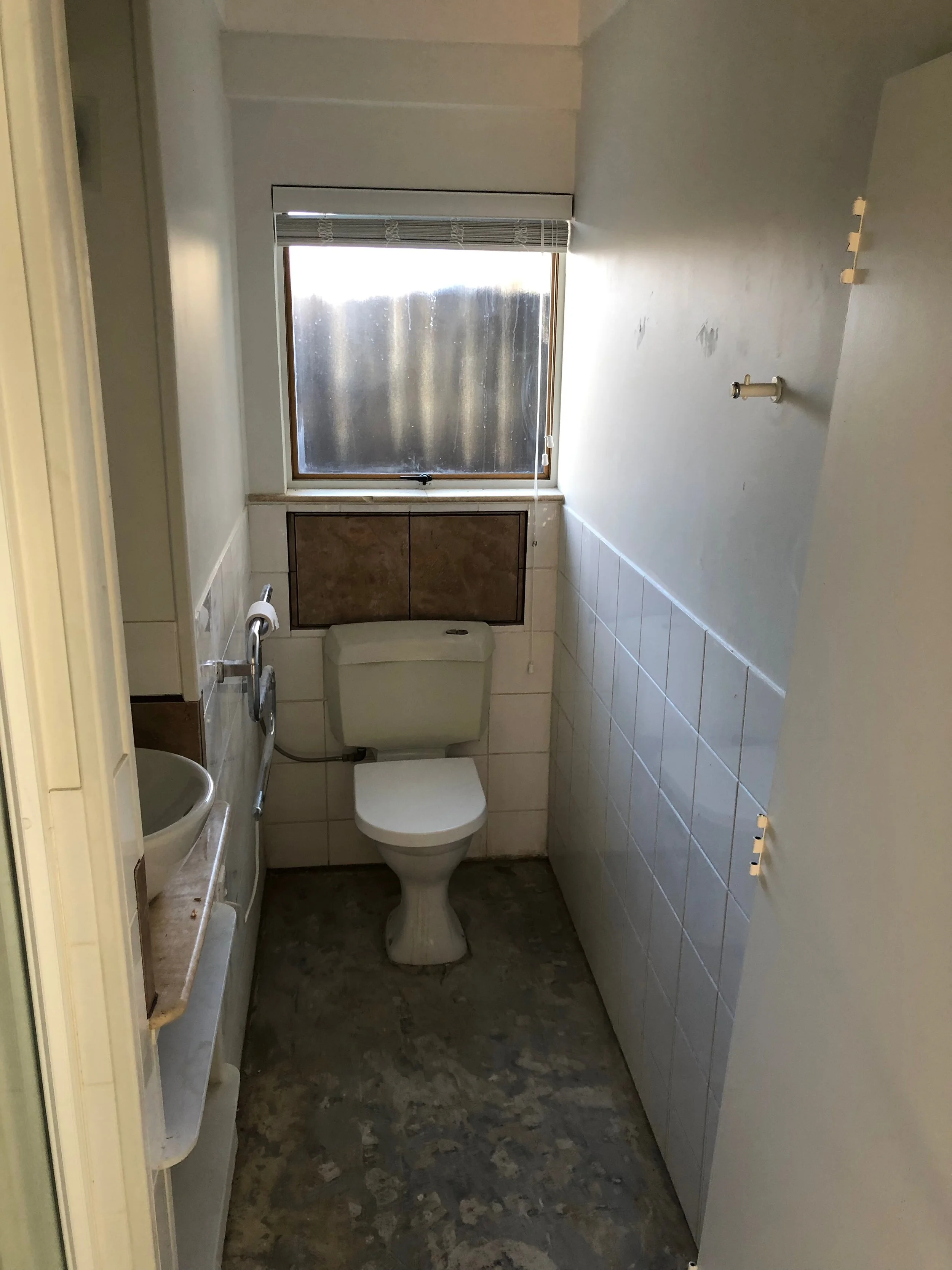 A small, unfinished bathroom with a toilet, a small sink on the left, and a window with partially closed blinds. The floor is bare concrete, and the walls are partially tiled and painted. There are some exposed fixtures on the wall to the right.
