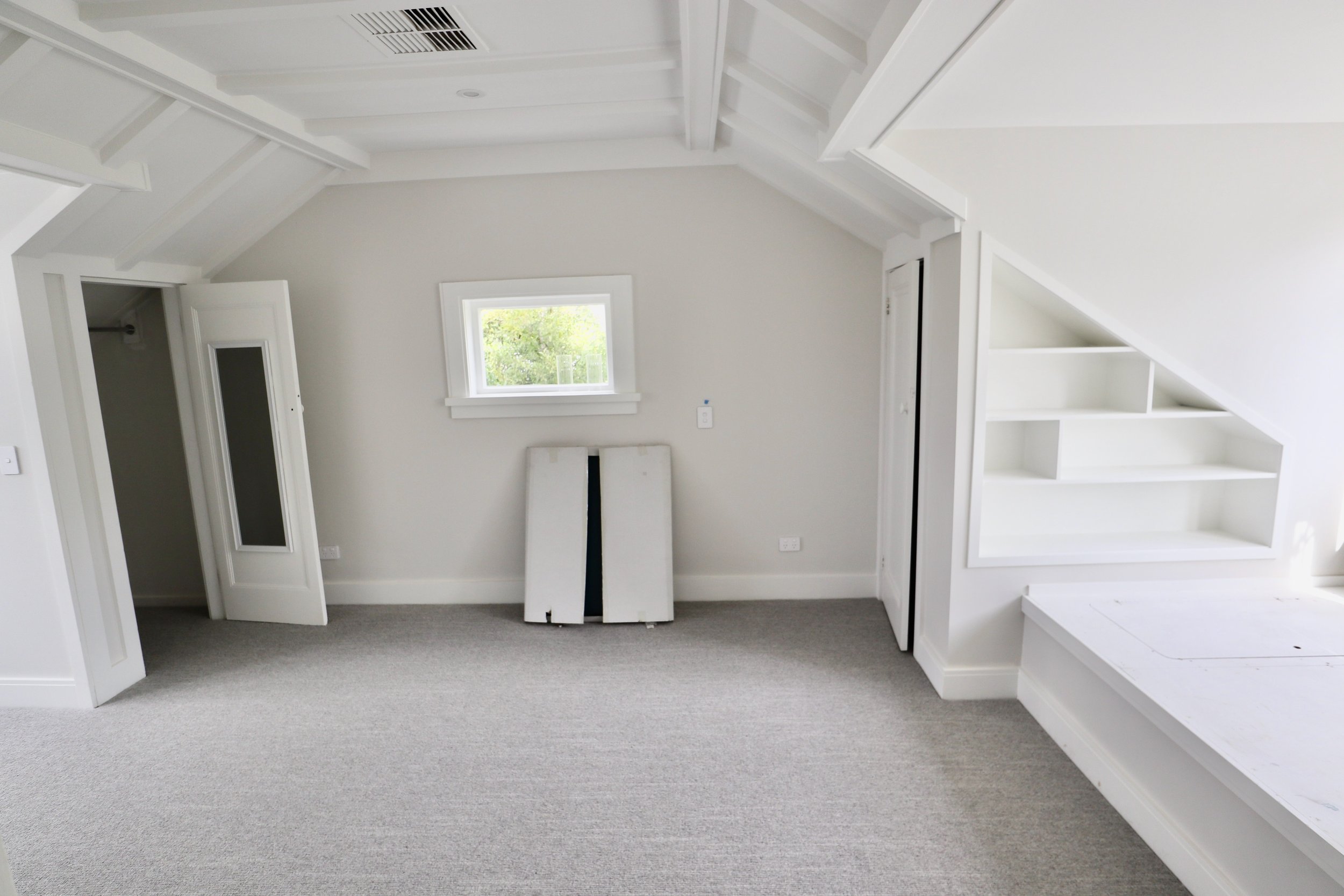 Empty white room with sloped ceiling, window, built-in shelves, grey carpet, and a folded bed frame against the wall.