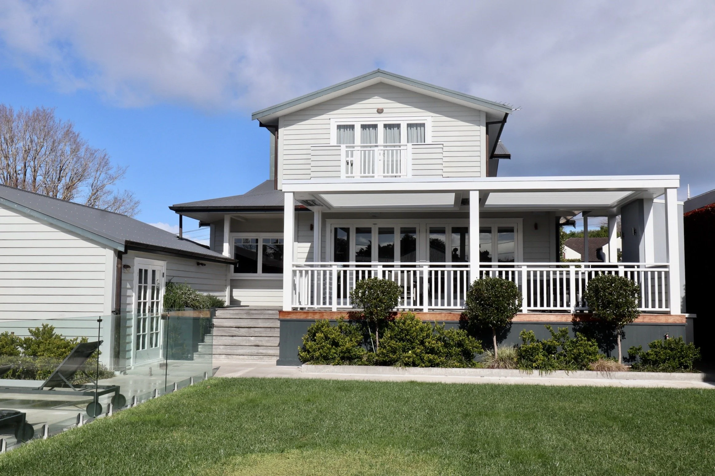 A two-story white house with a large porch and a balcony, surrounded by a lush green lawn and manicured bushes, under a partly cloudy sky.