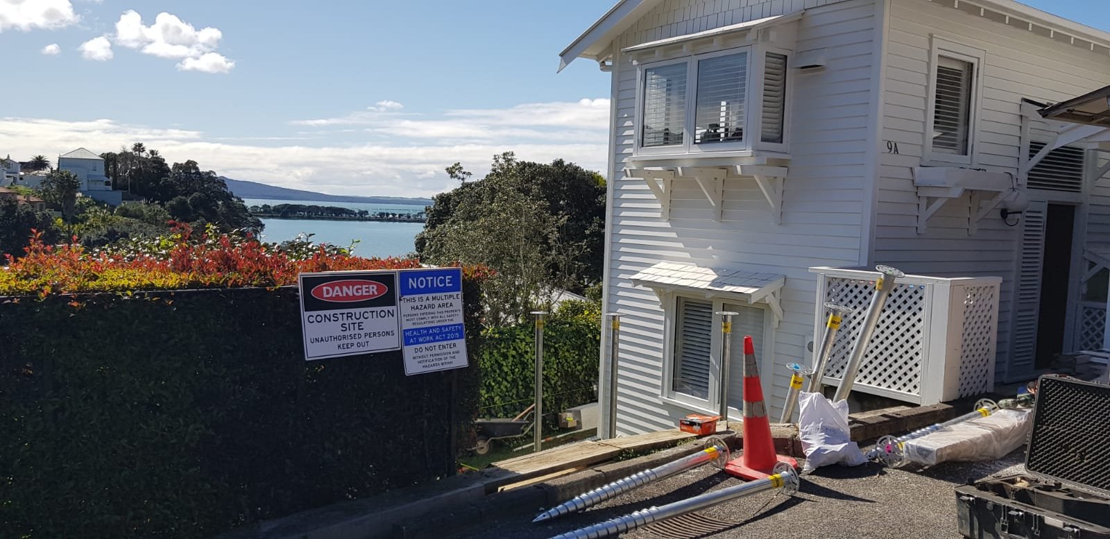 A construction site next to a white residential building with a ocean view in the background, warning signs on a fence, construction tools, and a traffic cone.