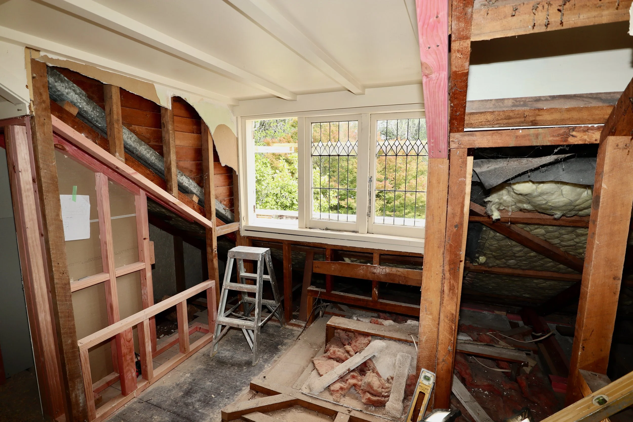 Interior view of a room under renovation, with exposed wooden framing, insulation, and a large window showing trees outside.
