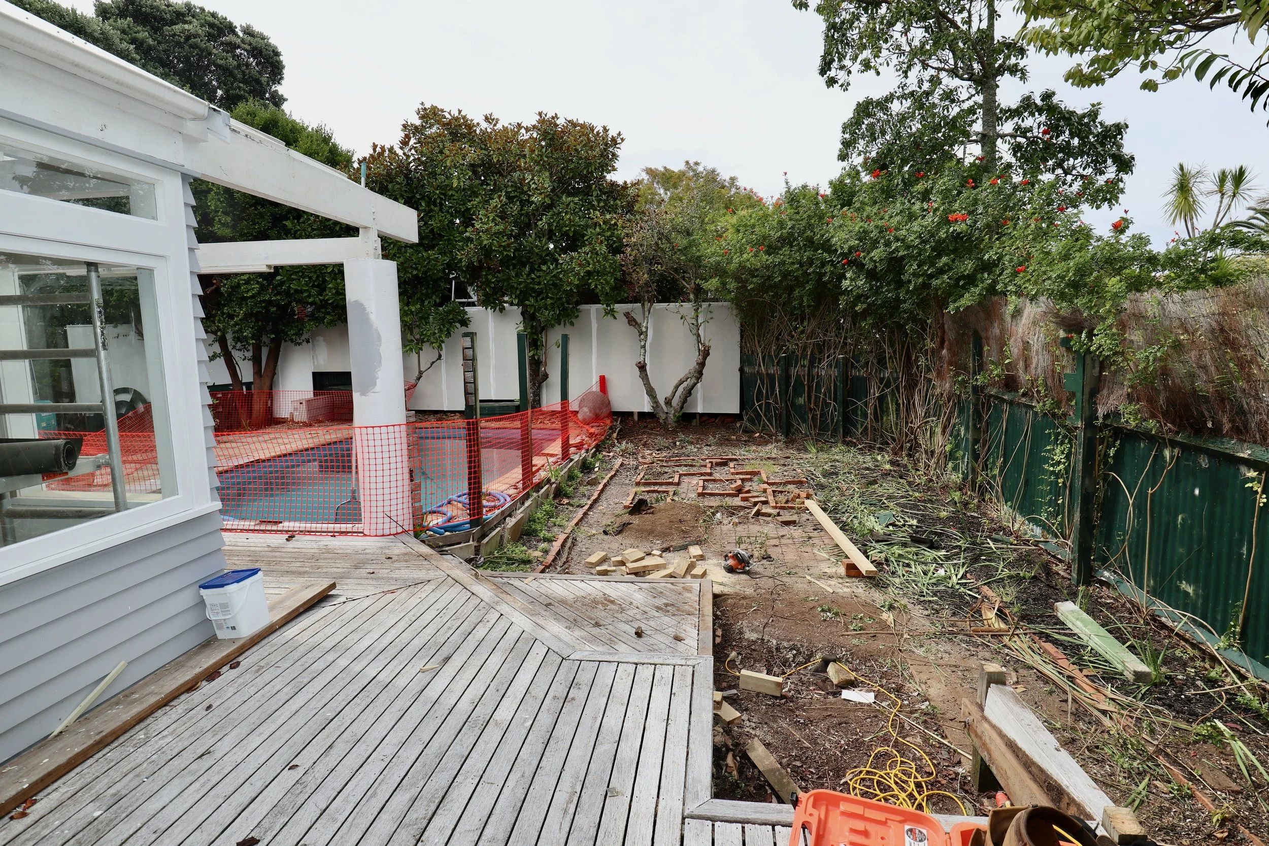 Backyard under renovation with a wooden deck, construction tools, and scattered bricks, trees, and a green fence along the yard's perimeter.