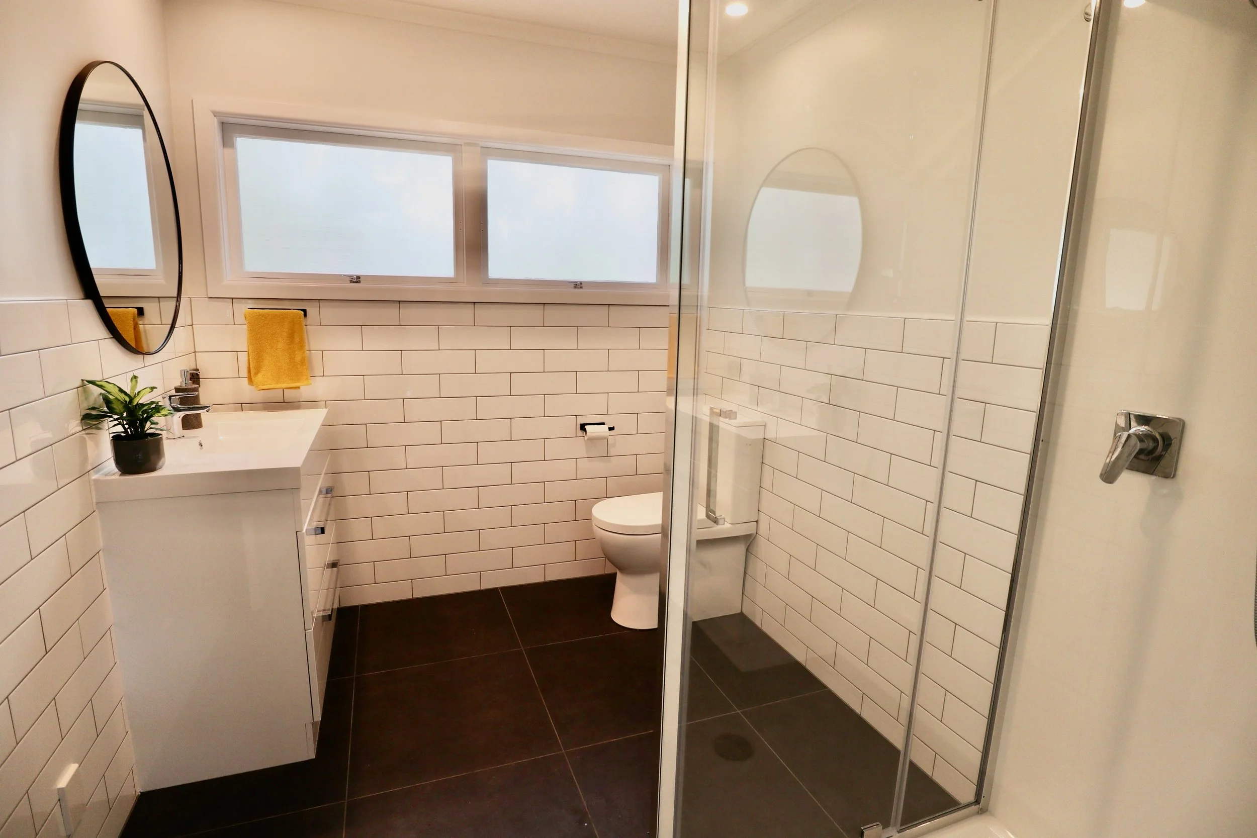 Modern bathroom with white subway tiles, dark floor tiles, a small white vanity with a black pot plant, yellow towels, a toilet, and a glass-enclosed shower area.