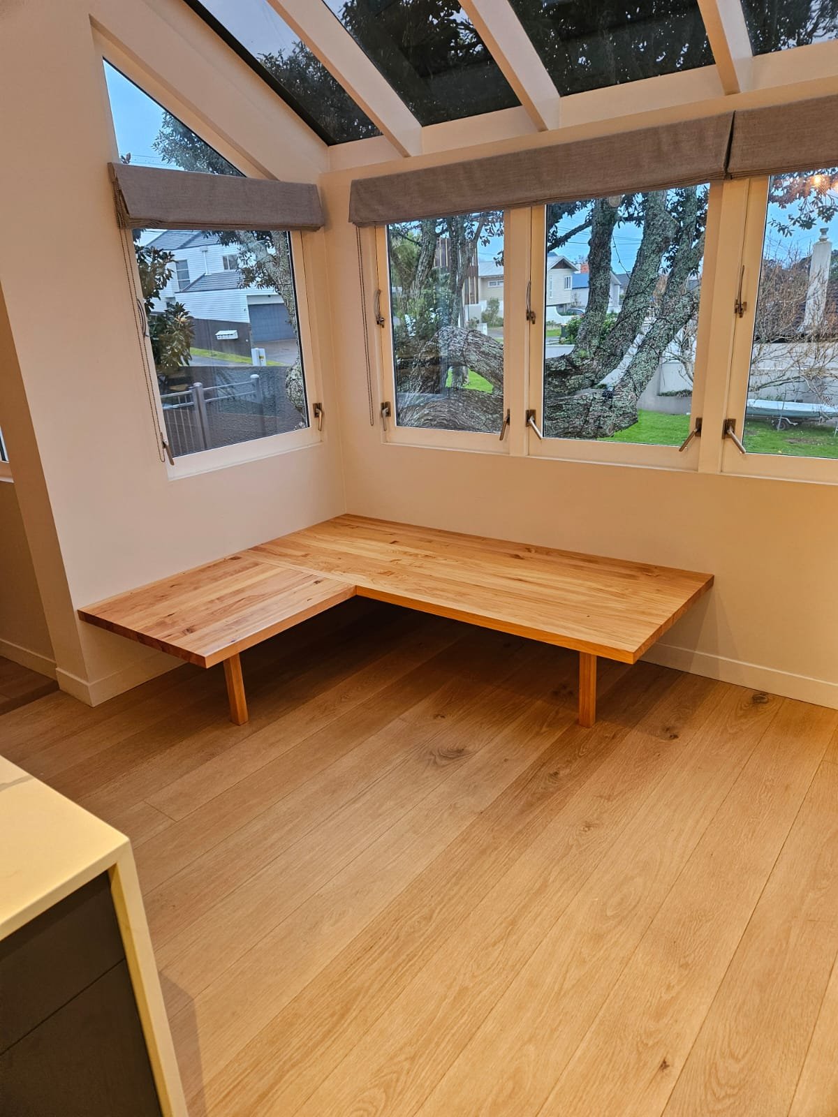 A room with large windows and a wooden floor, featuring a low wooden bench beneath the windows, with trees and houses visible outside.