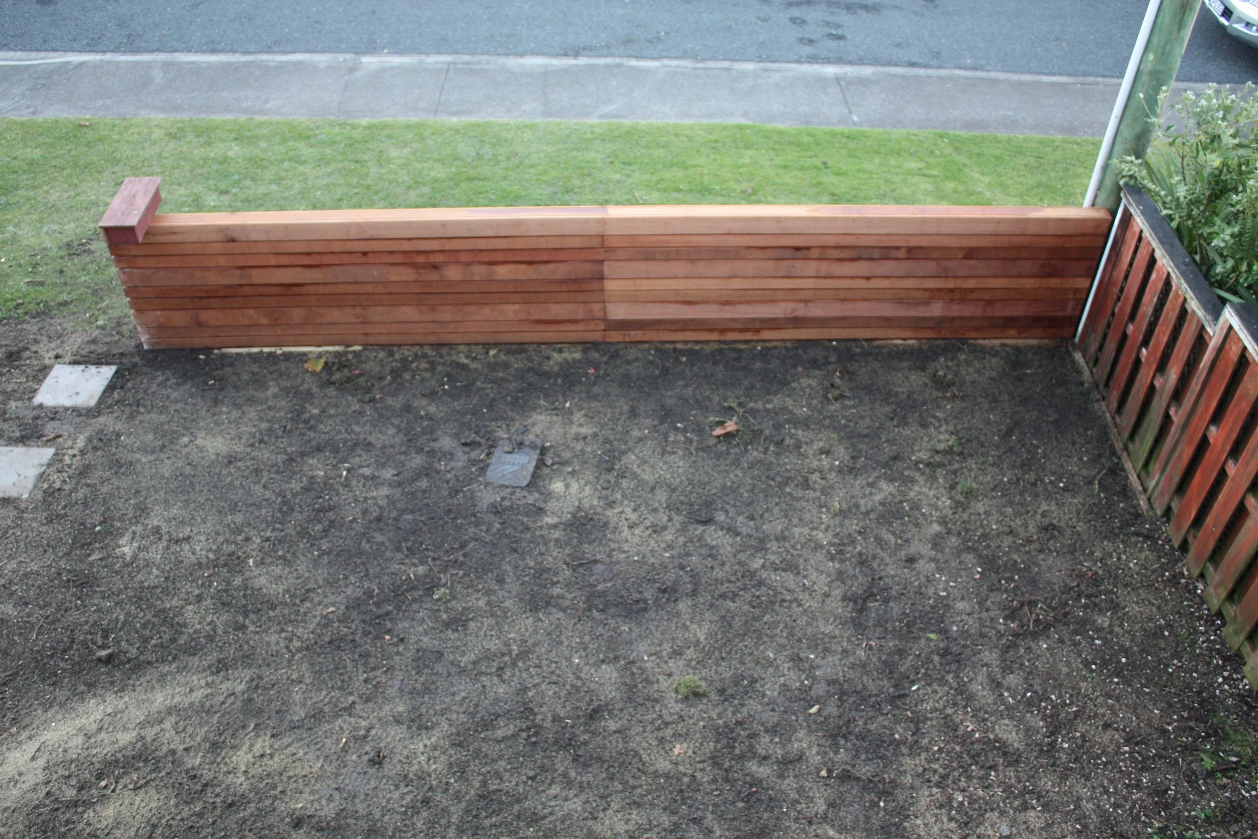 A newly built wooden fence with horizontal planks in a small backyard, with a patch of dirt in front and a grassy lawn beyond, with a street and parked cars in the background.