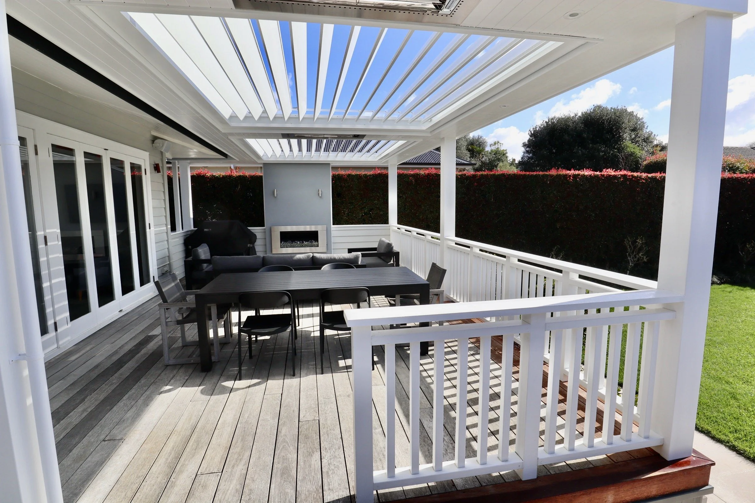 Outdoor porch with black dining table, black chairs, a grey couch, and a wall-mounted fireplace surrounded by white railings and a pergola ceiling, with a grass lawn and hedge in the background.