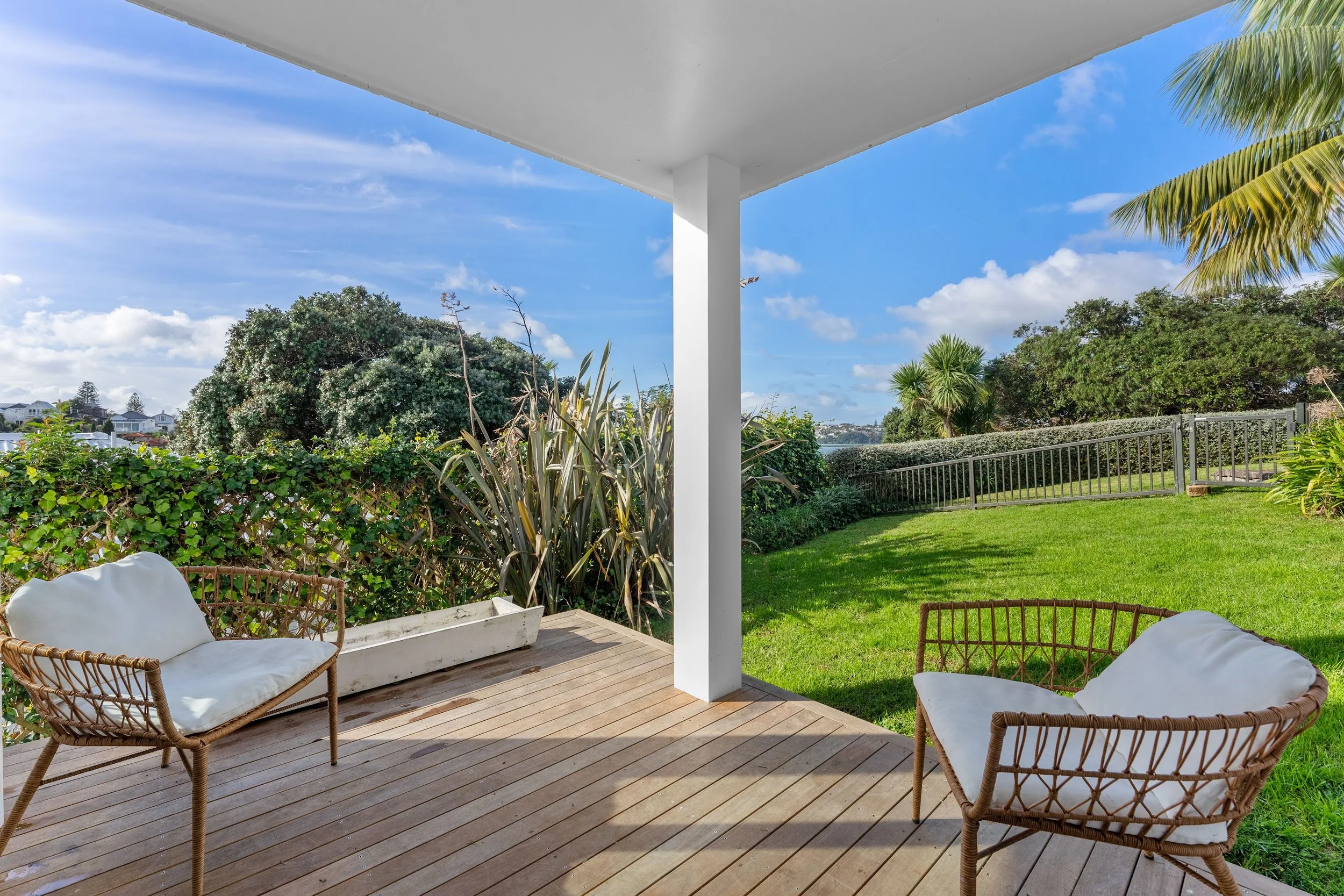 View of a backyard patio with two rattan chairs with white cushions, a white planter box, green grass, various trees and bushes, a metal fence, blue sky with clouds, and a distant view of water and houses.