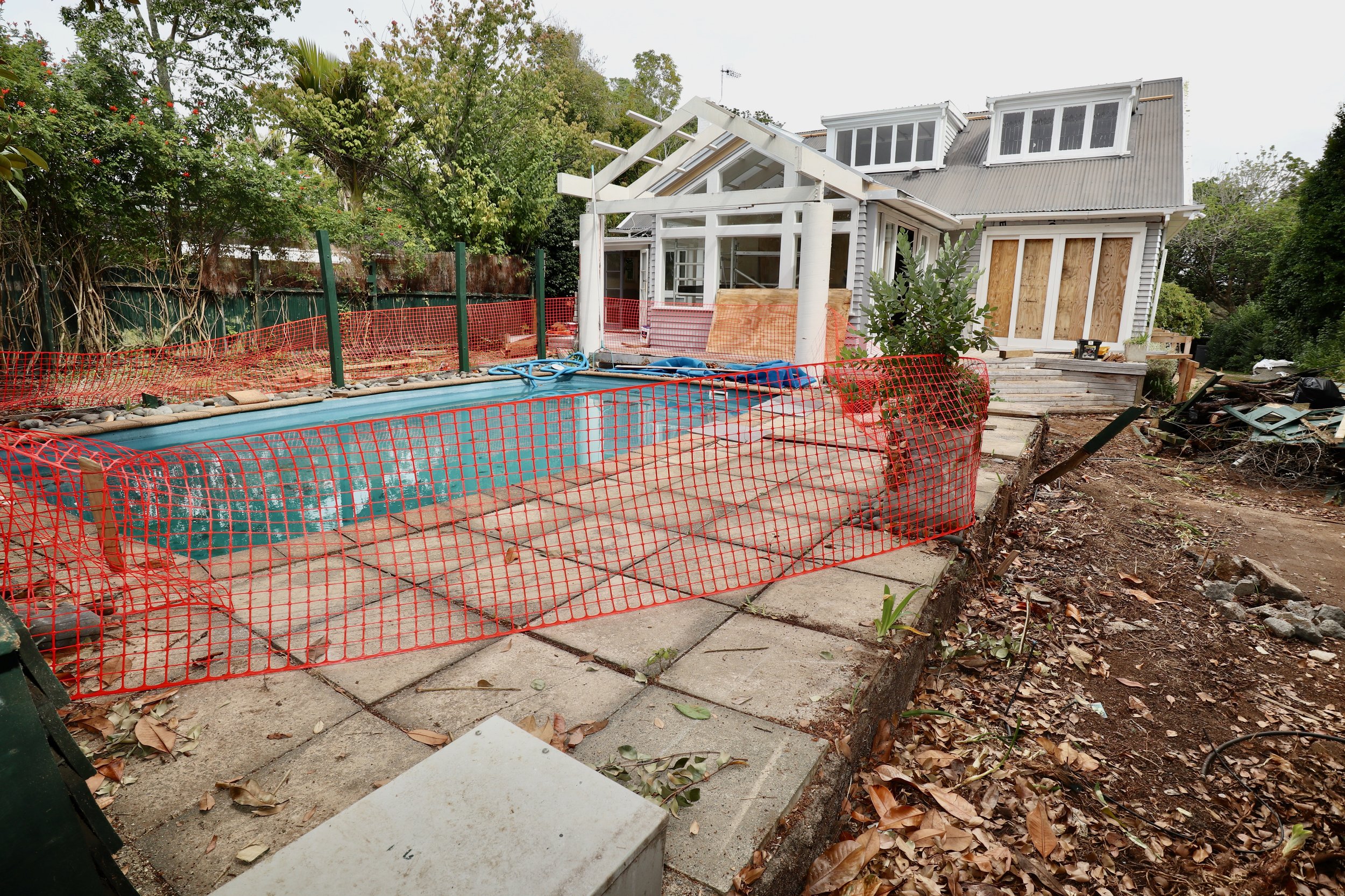 A backyard with a swimming pool surrounded by a construction site. The pool is fenced off with orange safety netting and construction materials are visible around it. There is a house in the background with some windows and an unfinished porch.