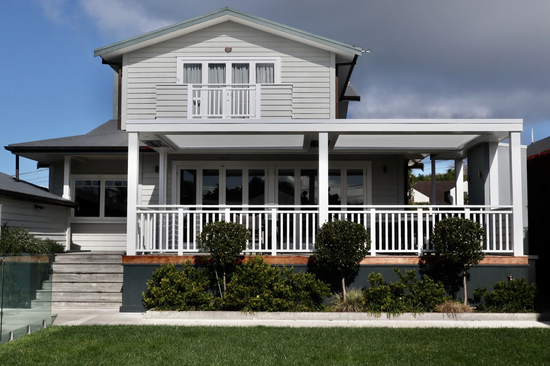 A two-story house with a white exterior, balcony on the upper level, and a covered porch on the lower level. The house has a gabled roof, large windows, and a front yard with small bushes and a grassy lawn.