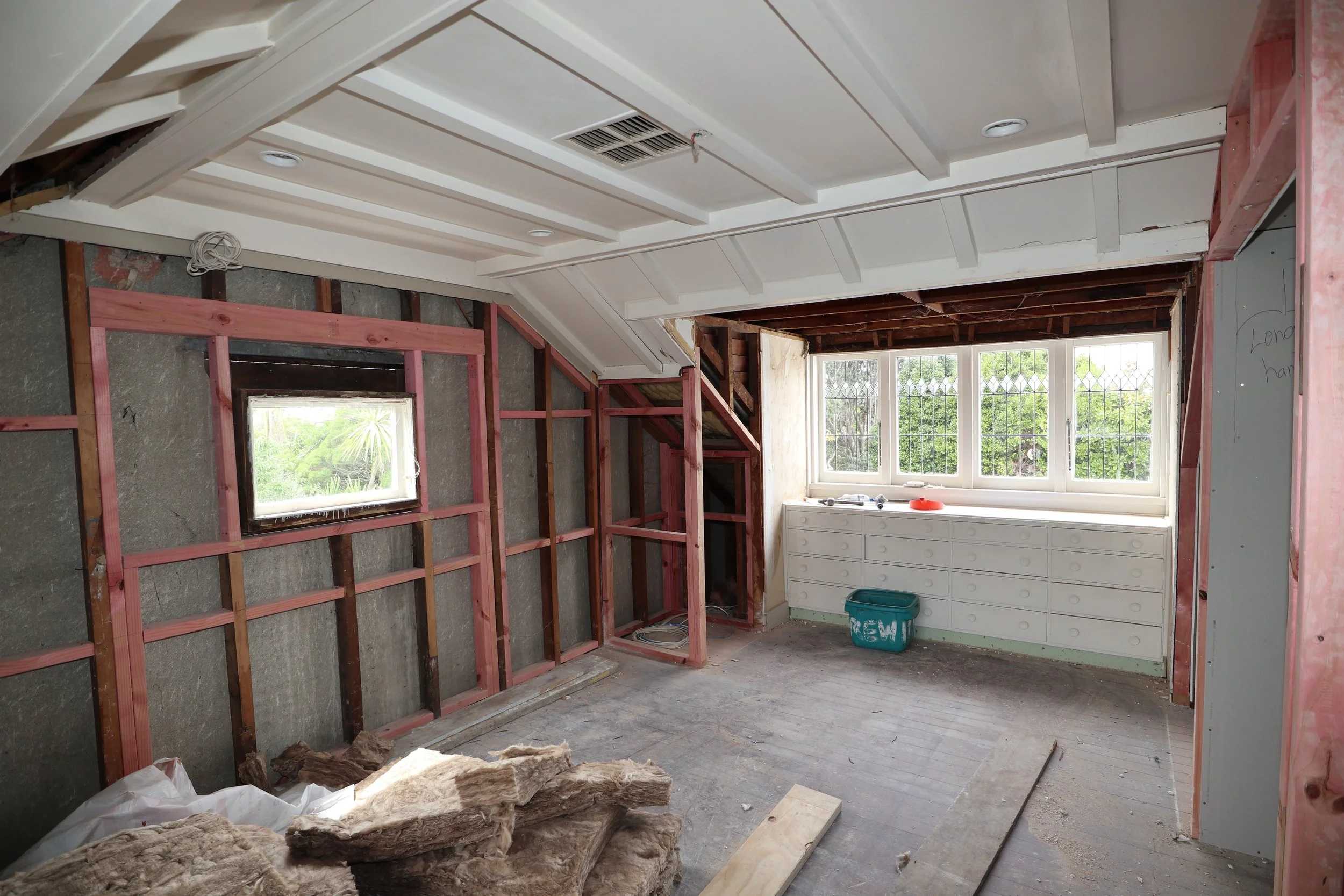 Room under renovation with exposed framing on the walls and ceiling, a window on the far wall, and construction materials on the floor.