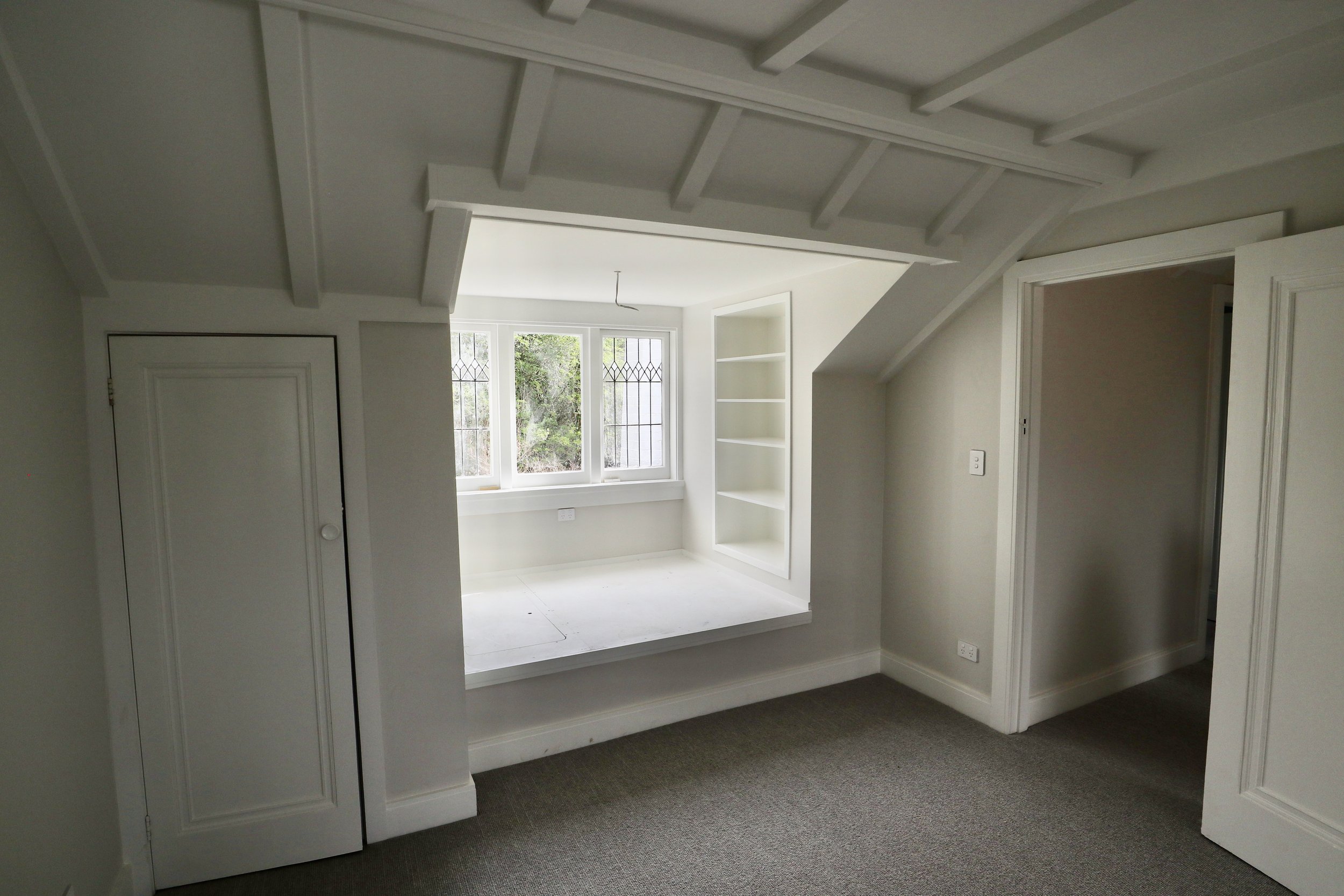 Empty room with a window seat, built-in shelves, and a sloped ceiling with exposed beams, painted white, with neutral carpeting.
