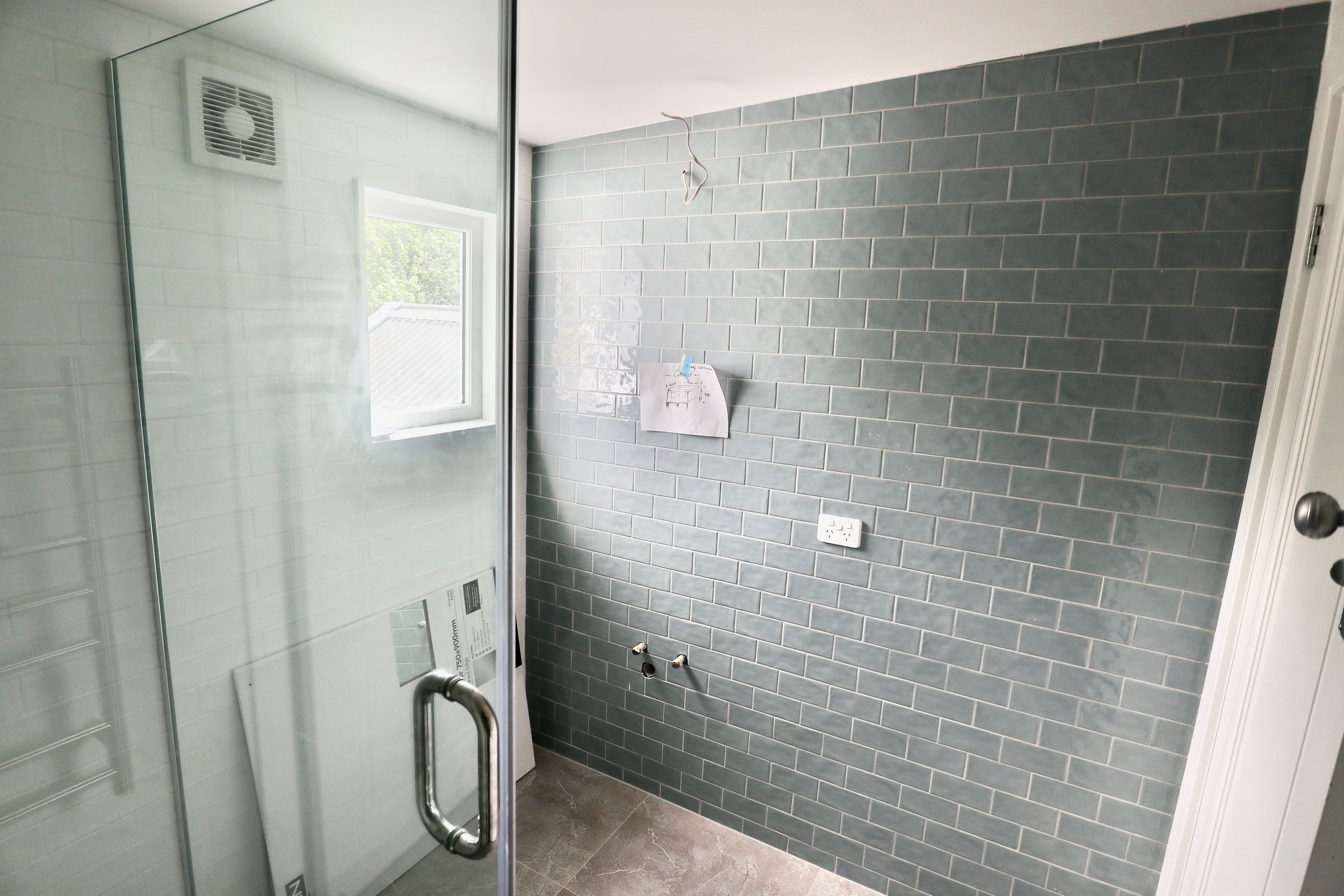 Bathroom with grey subway tile walls, a large window, and a glass shower enclosure. There are exposed plumbing connections and electrical outlets on the wall.