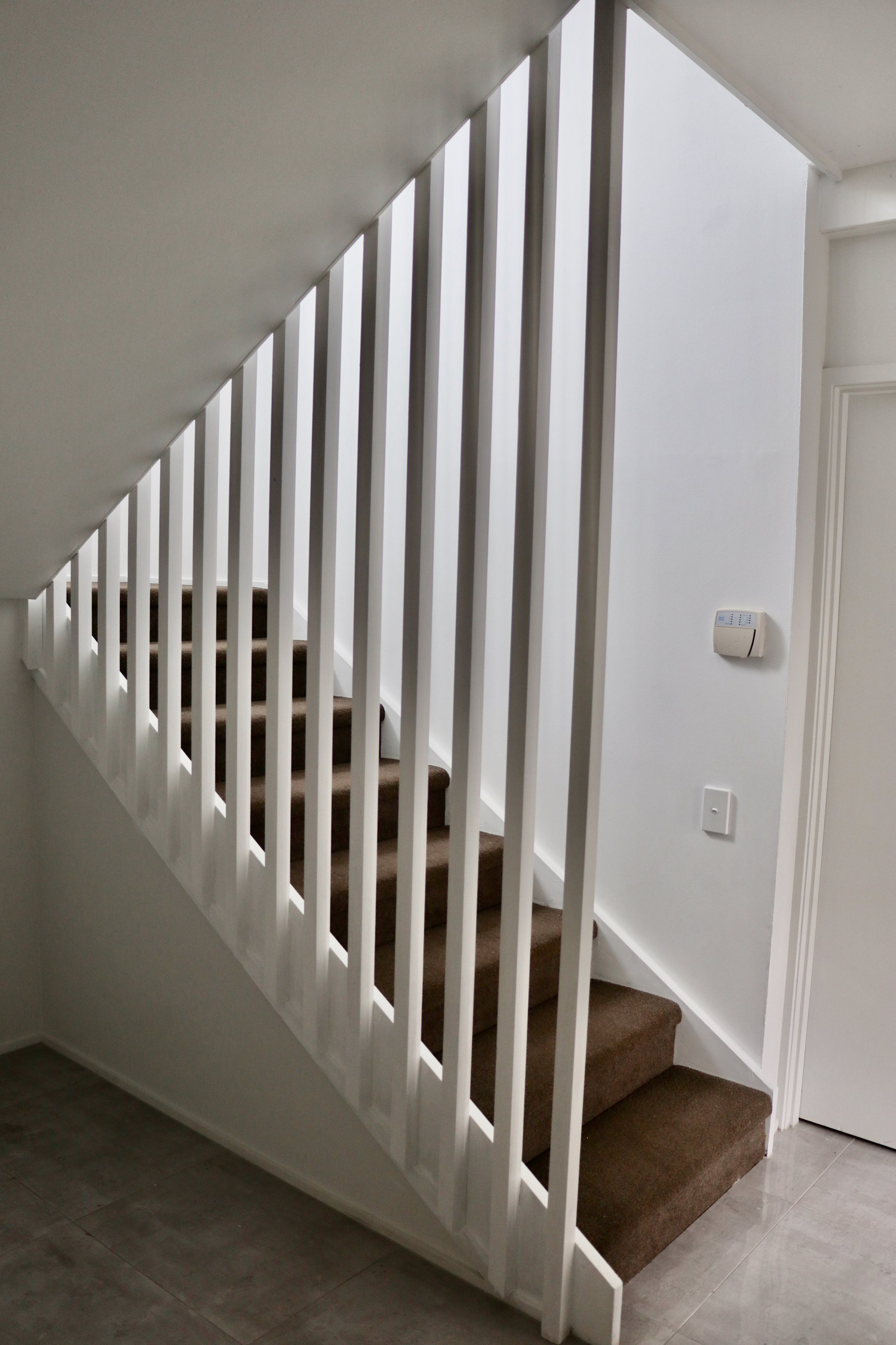 Interior view of a staircase with beige carpeted stairs and white railings, next to a white wall with a thermostat and a door.