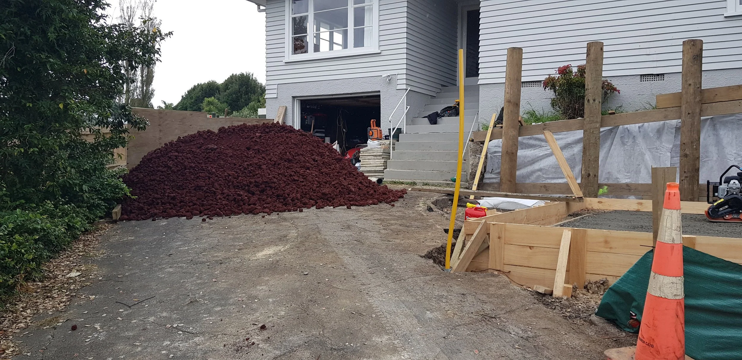 Construction site in front of house with orange traffic cone, wooden framing, and a pile of dirt or mulch.