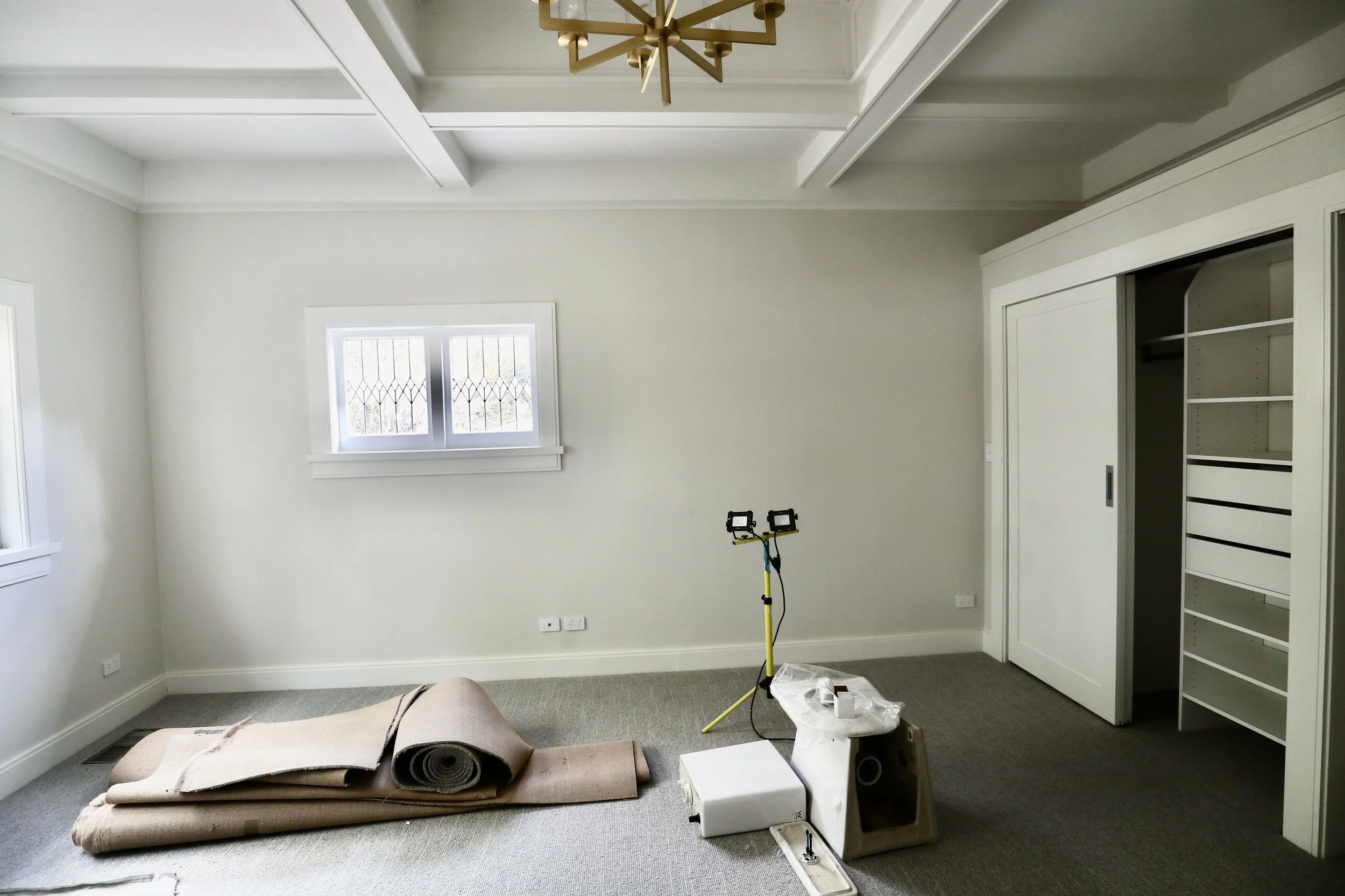 Empty room with light-colored walls, carpeted floor, two small windows, and a built-in closet. Construction tools and materials are placed on the floor, indicating ongoing renovations.